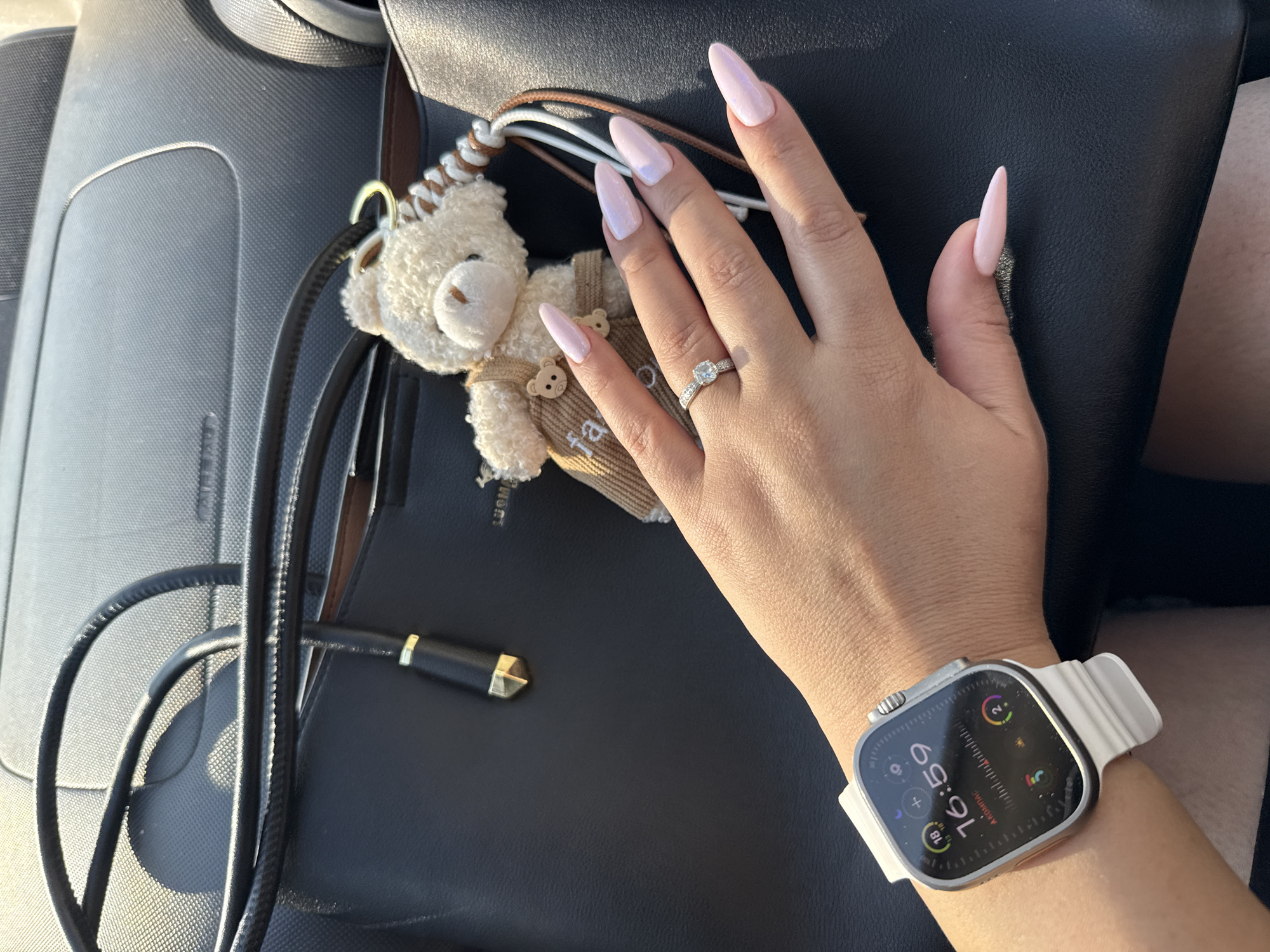 The image shows a close-up of a person's left hand, adorned with a white Apple Watch and a ring on their ring finger. The hand has long, light pink, almond-shaped acrylic nails. The person is wearing the watch in a car, indicated by the dashboard visible in the background and the texture of the car seat. A black purse with a teddy bear keychain is also visible, suggesting the person is likely on the go. The lighting indicates it is daytime, with sunlight casting shadows. There is no indication of the location being in Tallinn, Estonia.