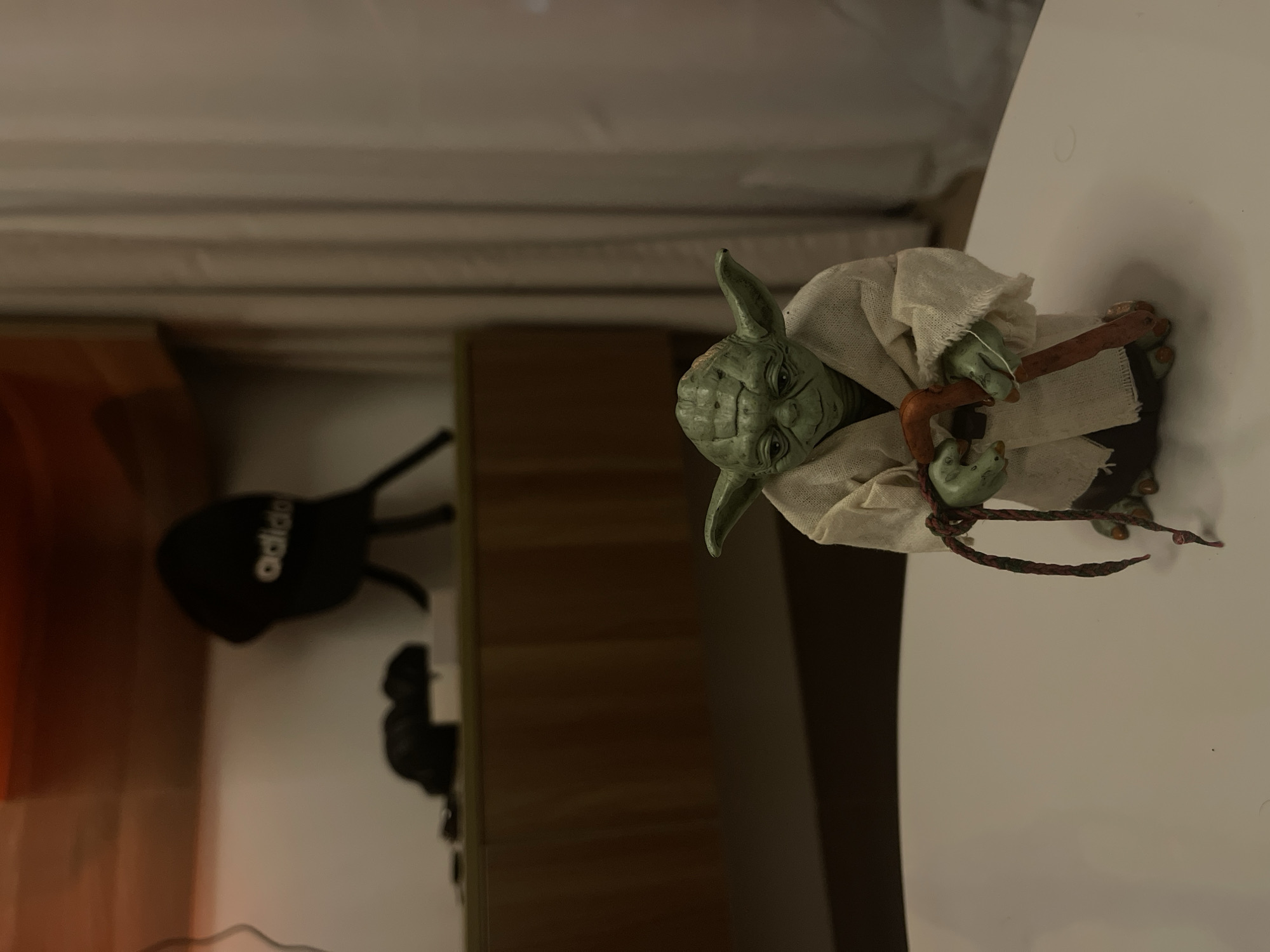 An action figure of the character Yoda from Star Wars stands on a white table, holding a brown cane. Yoda is depicted wearing a tan robe and has green skin and large pointed ears. The figure is in focus, while the background is blurred. In the blurred background, a shelf with some items, including a black baseball cap with "adidas" written on it, is visible. A beige curtain is also present in the background. The lighting in the scene is dim, with a warm tone. The image does not provide any information about a specific location in Đà Nẵng, Vietnam.