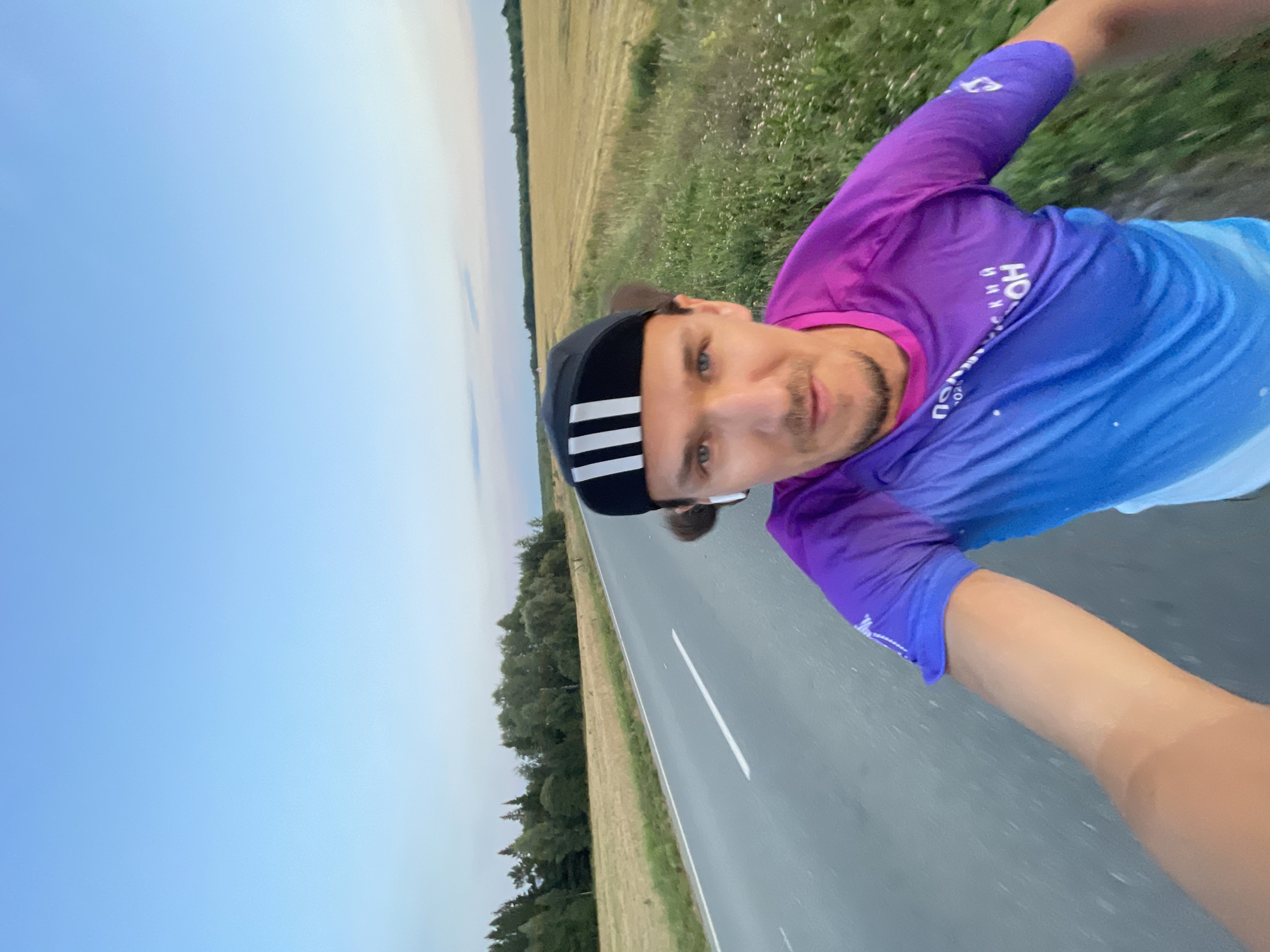 In this selfie-style shot, a man wearing a black cap with white stripes and a purple and blue gradient t-shirt with Cyrillic text on it is captured. He is outdoors on a paved road, with a field of dry grass and a line of trees in the background. The sky is a clear blue with a hint of sunset colors. He has earbuds in his ears and appears to be in motion, likely running or cycling given the dynamic angle of the shot and the background blur. The location is likely a rural area in Russia.