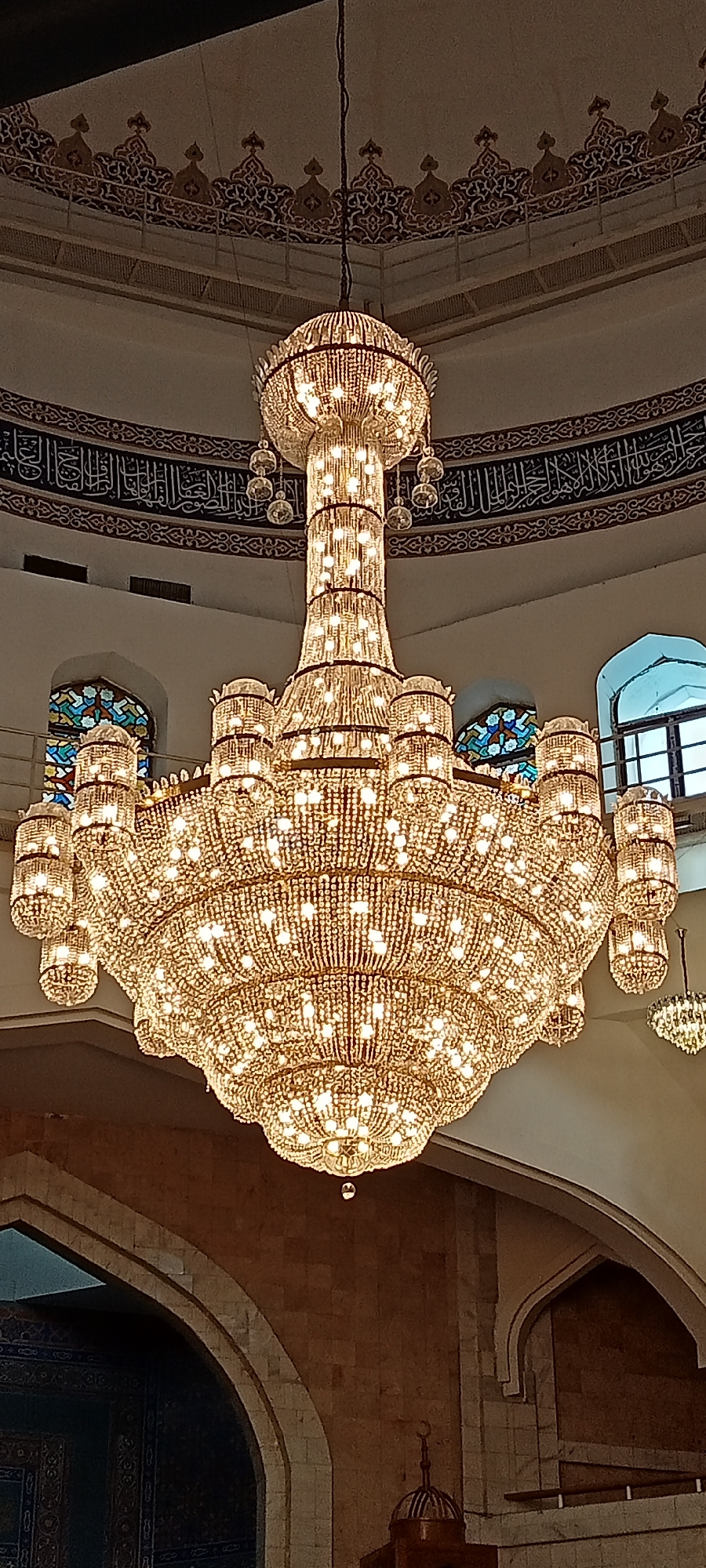 A large, ornate crystal chandelier hangs from the ceiling of a building, illuminating the interior. The chandelier is multi-tiered and densely covered in crystals, with numerous lights creating a dazzling effect. The ceiling features decorative Islamic calligraphy and architectural elements, including arched doorways and stained-glass windows visible in the background. The walls appear to be constructed of stone or brick. The overall scene suggests an interior of a mosque or a grand religious building. The image's metadata indicates it was taken in Almaty, Kazakhstan.