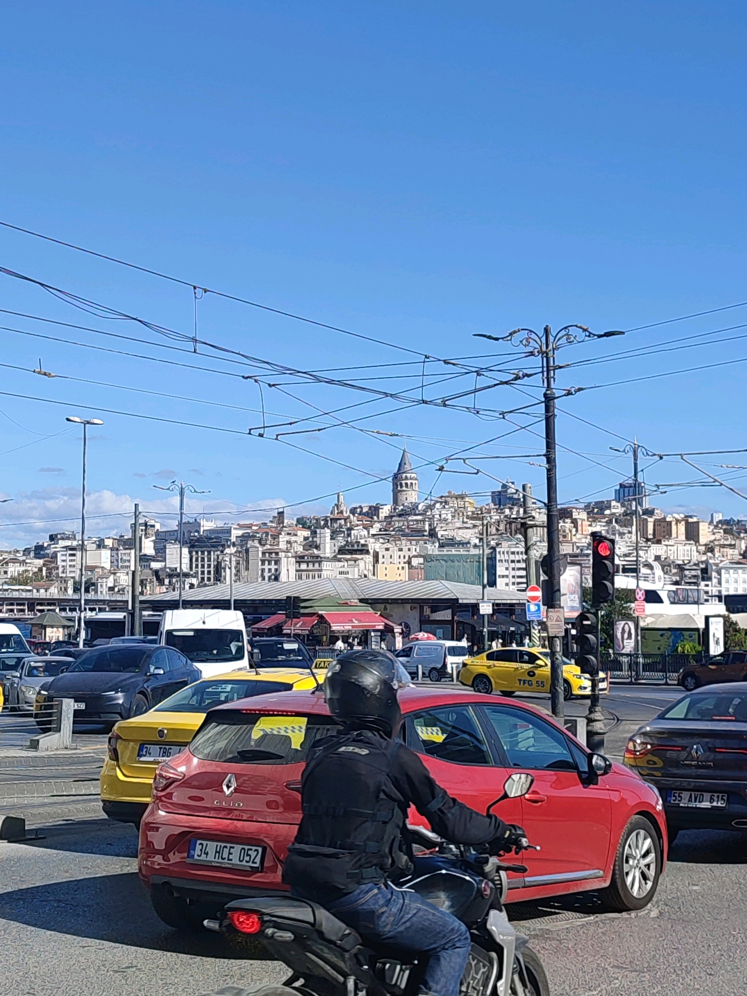 This is a vertical, eye-level, outdoor shot of traffic in Istanbul, Turkey, featuring the Galata Tower in the distance. The foreground is dominated by a motorcycle with a rider wearing a black helmet and jacket, in the process of navigating through traffic. In front of the motorcycle is a red Renault Clio with a license plate that reads "34 HCE 052." To the left of the Clio is a yellow taxi with the license plate "34 TBG 599." Several other cars and a white van are also visible, creating a bustling urban scene. Overhead, a complex network of electrical wires crisscrosses the sky. The background showcases the cityscape of Istanbul, characterized by densely packed buildings and the iconic Galata Tower. A traffic light is visible, showing a red signal. The overall scene suggests a busy street in Istanbul on a clear, sunny day.