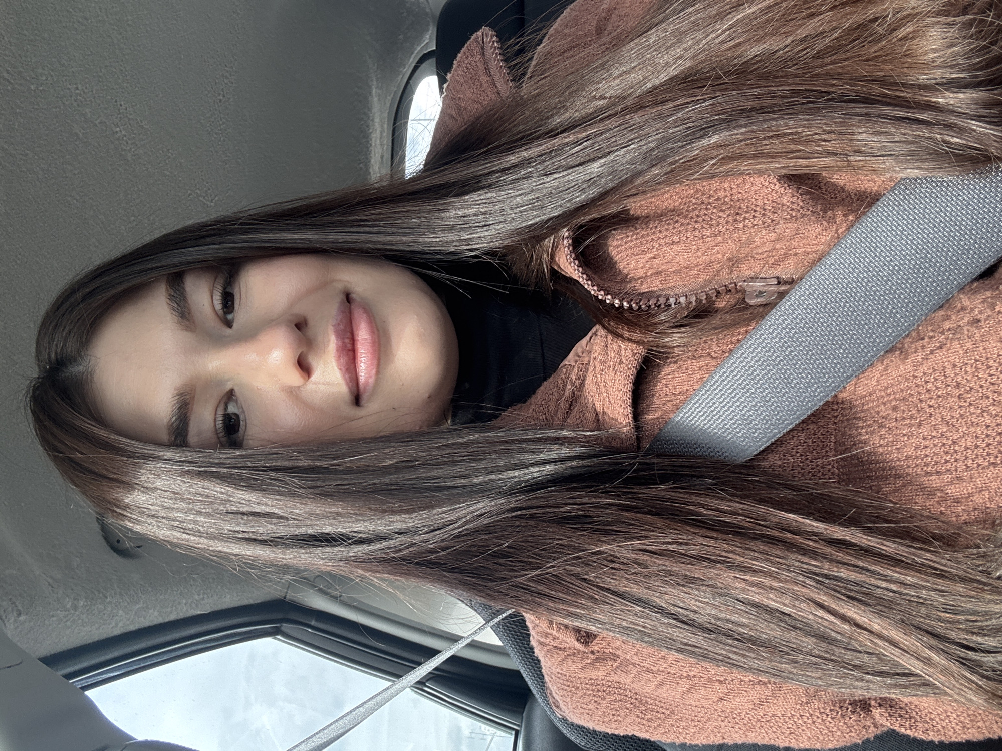 A woman with long brown hair is sitting in a car, wearing a brown jacket and a seatbelt. The image appears to be taken from a low angle, looking upwards. The location cannot be determined as the image does not contain information about the city of Chaikovsky, Russia.