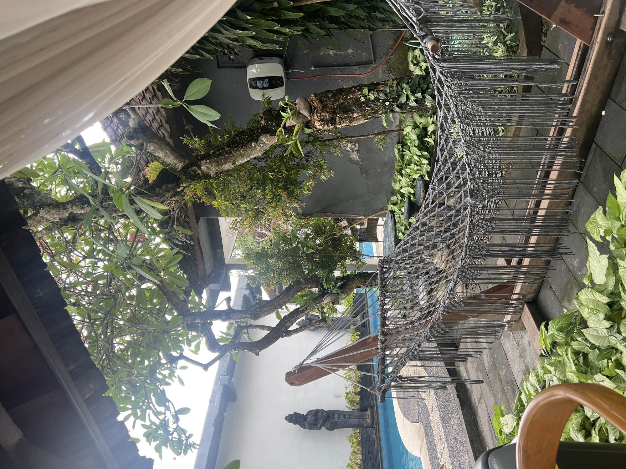 This sideways-oriented image captures a serene outdoor tropical setting, likely a garden or resort area in Kuta Selatan, Indonesia. The scene is bathed in bright, diffused daylight, suggesting an overcast day or shaded spot.
Prominently featured are several large trees with thick, gnarled trunks and lush green canopies, some appearing to be Frangipani with their distinctive leaves. Smaller plants and moss adorn the tree trunks, contributing to the rich, natural environment. On a dark grey wall behind one of the trees, a modern white, wall-mounted water heater with a blue digital display and a red connecting cable is visible.
To the right, a large, dark grey macrame or rope hammock is suspended, featuring an intricate woven pattern and numerous long tassels that hang downwards. It appears to be supported by a sturdy wooden beam structure.
In the lower part of the scene, a blue-tiled swimming pool is visible, edged with decorative mosaic-patterned stone work. Next to the pool, against a pristine white wall, stands a dark stone statue, resembling a traditional Balinese deity or figure. The surrounding ground is covered with various vibrant green plants, including some with variegated leaves.
Architectural elements such as parts of dark-tiled rooflines are visible in the upper and lower sections of the frame, hinting at nearby buildings. A partial view of a wooden chair arm can be seen in the bottom right corner. The overall impression is one of tranquility and natural beauty, characteristic of a tropical retreat. No legible text is discernible other than blurry symbols on the water heater's display.
