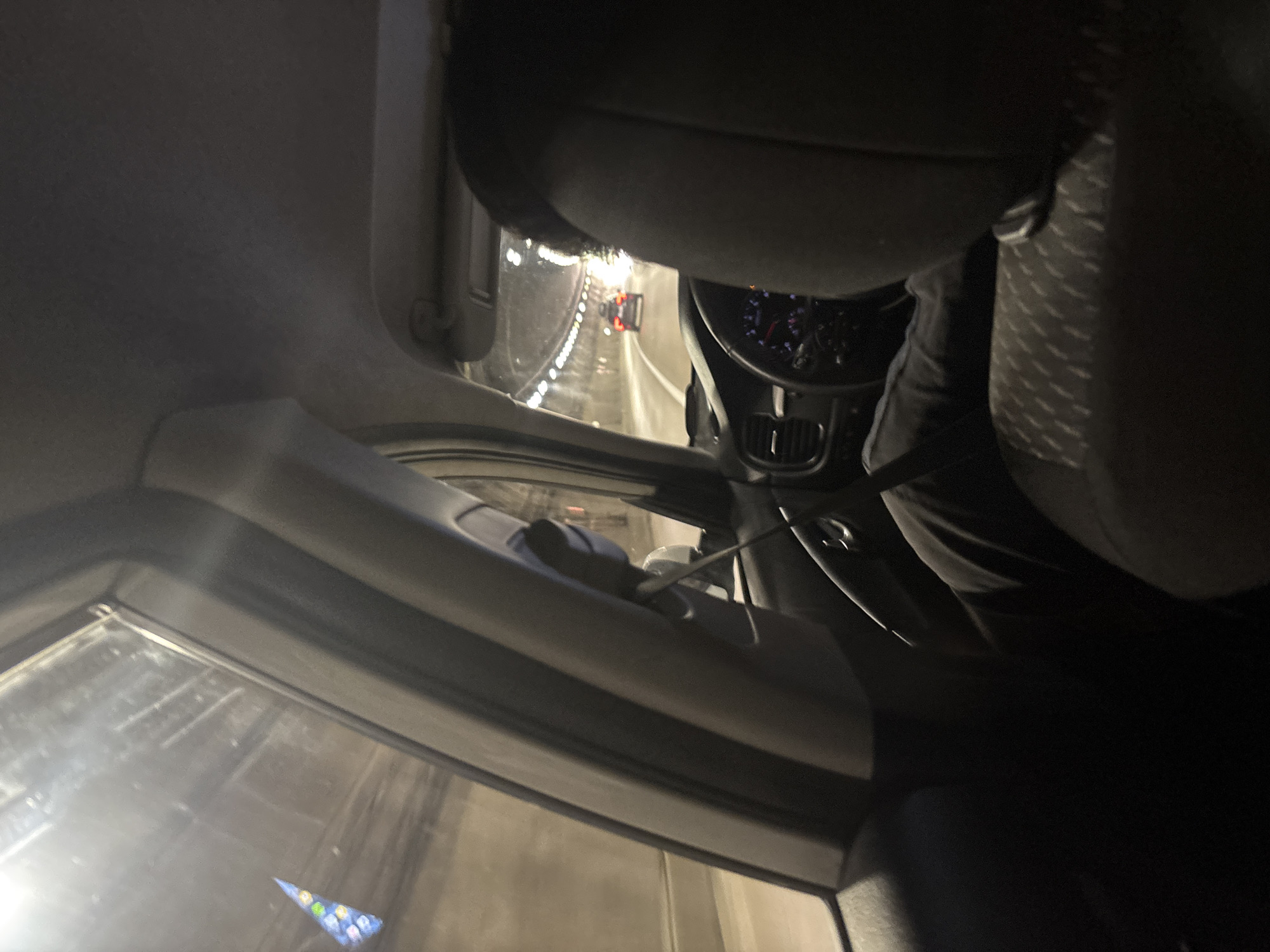 The image shows the interior of a car driving through a tunnel. The perspective is from the back seat, looking towards the front. A person's head and shoulders are visible in the foreground, seated in the driver's position. The steering wheel, dashboard, and part of the driver's legs are also visible. The tunnel is illuminated by overhead lights, and another car is visible ahead in the same lane. The exterior of the tunnel walls is visible through the side windows. There is no indication of the city of Kepsha, Russia, in the image.