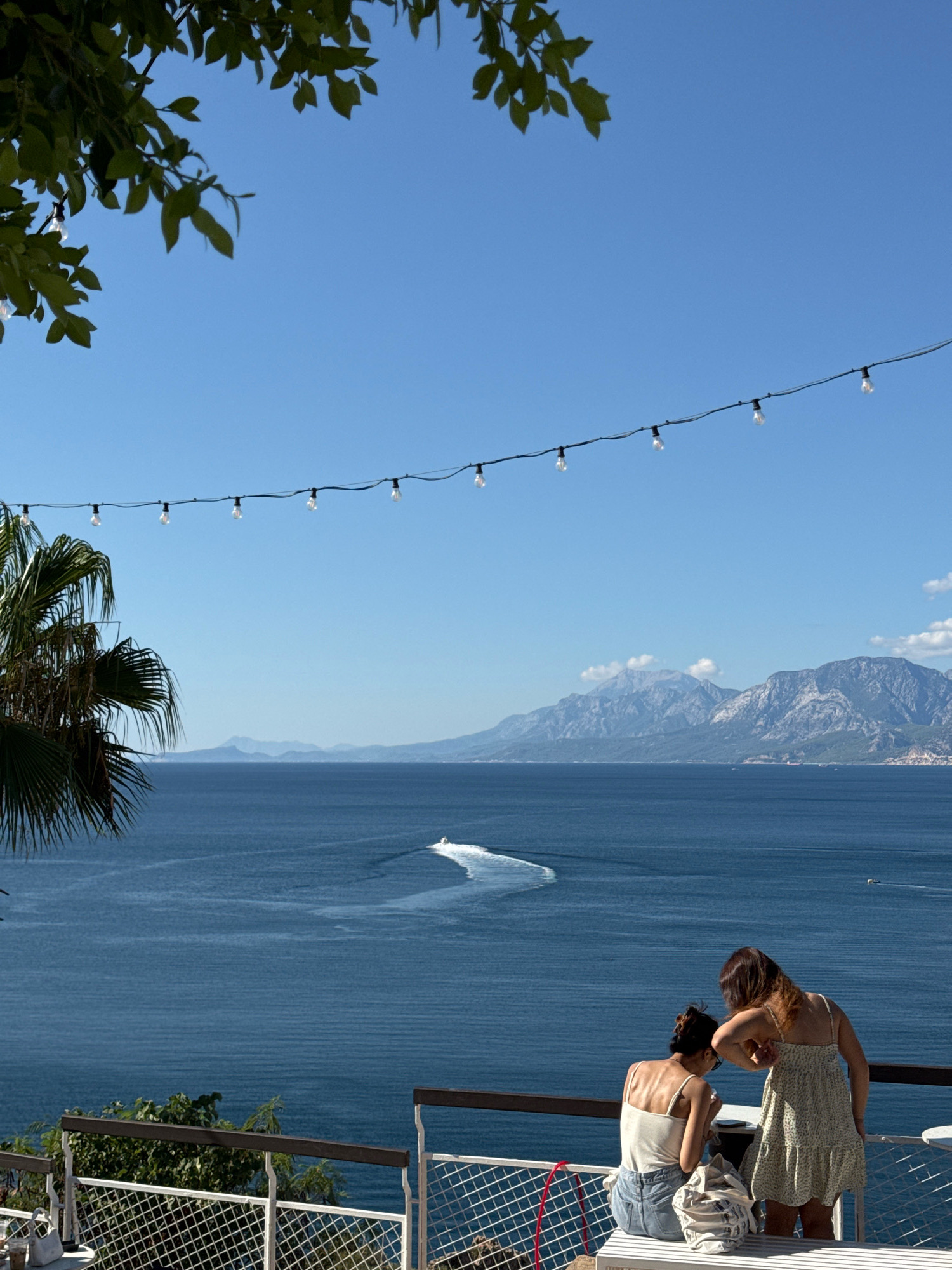 Two women are seated at a white bench on a balcony overlooking the Mediterranean Sea. The woman on the left is wearing a white tank top and denim shorts. The woman on the right is wearing a floral print dress. Both women are looking down, engaged in conversation. A white tote bag rests on the bench between them. A string of lights stretches across the top of the frame. In the distance, mountains rise from the coastline. A speedboat creates a white wake on the deep blue water. Lush green foliage frames the top left corner of the image, and a palm frond is visible on the left side. The sky is clear and blue. This view is characteristic of coastal areas in Antalya, Turkey, known for its scenic beauty and seaside resorts.
