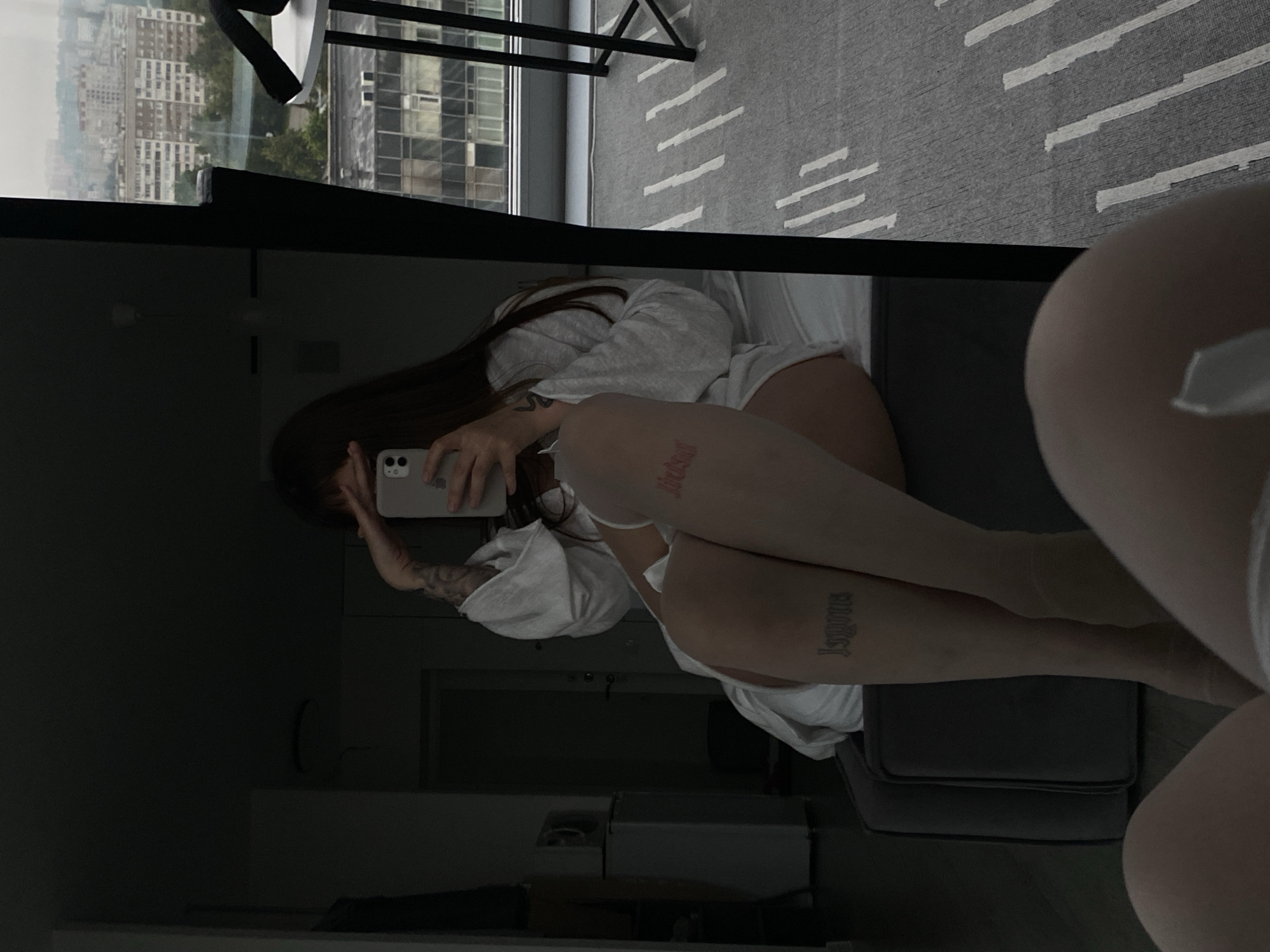 A woman is reflected in a mirror in an urban apartment. She is wearing a white robe and beige tights, with tattoos on her legs. One tattoo on her right thigh reads "impure," and another on her left thigh is red and appears to say "live." She is holding a phone and appears to be taking a selfie. The room is dimly lit, with furniture and a refrigerator visible. Outside the window, the city of Izhevsk, Russia, can be seen, with buildings and a cloudy sky. A table and chairs are also visible in the reflection.