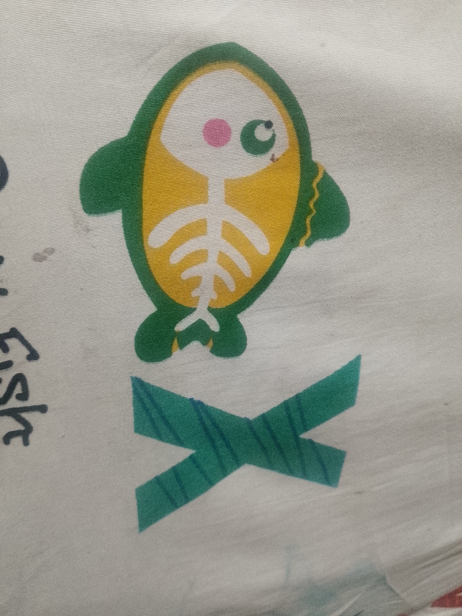 The image shows a portion of patterned fabric with a cartoon fish design. The fish is stylized with a green outline, a yellow body, a white skeletal structure, a pink cheek, and a green eye. Below the fish, there is a green "X" shape. A portion of the word "fish" is visible to the left of the fish illustration. The fabric appears to be white. The scene is not identifiable as Lhokseumawe, Indonesia. No people are present.