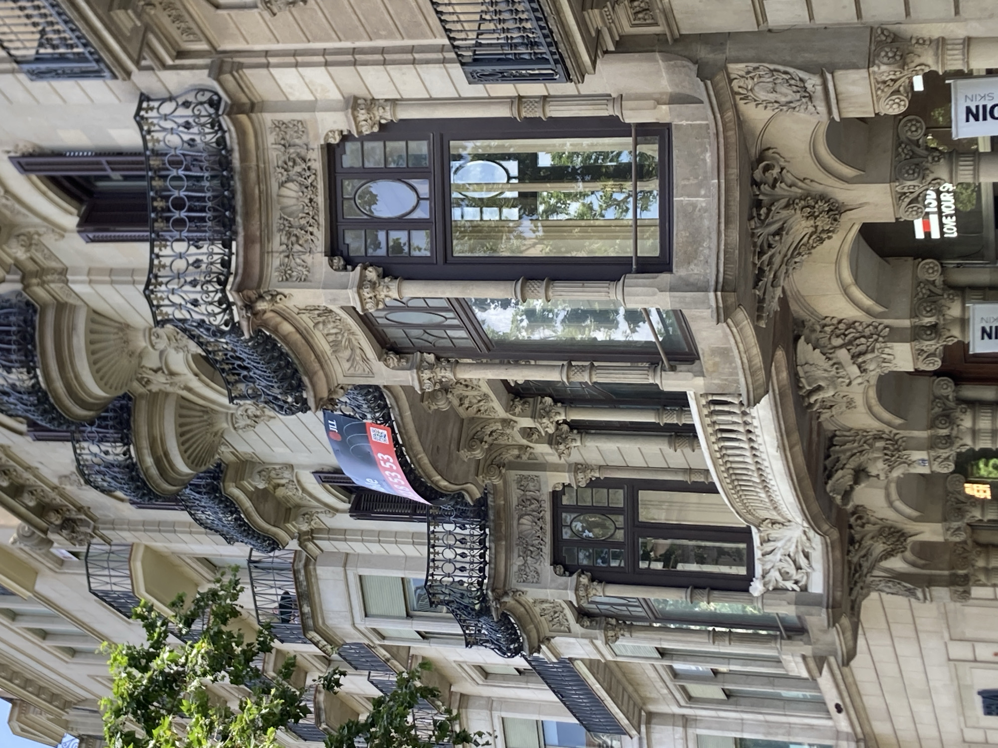 The image captures a close-up, eye-level view of the ornate facade of a building in Barcelona, Spain. The building's architecture is characterized by detailed stonework, curved balconies with intricate wrought-iron railings, and large windows. The main focus is on a prominent bay window section with decorative carvings at its base. Above the window, a balcony displays a red and blue banner with white text, likely an advertisement, featuring the JLL logo and a phone number. The building appears to be made of light-colored stone, with darker window frames and dark shutters. Lush green foliage from a tree is visible in the upper left corner, partially obscuring some of the upper floors and balconies. The lighting suggests it is daytime, with sunlight reflecting off the windows. The lower part of the image shows the entrance area with columns and more decorative stone work, and partial signage for "KIN SKIN" and a slogan that reads "LOVE YOUR S". The overall impression is one of elegant, historical architecture in an urban setting.