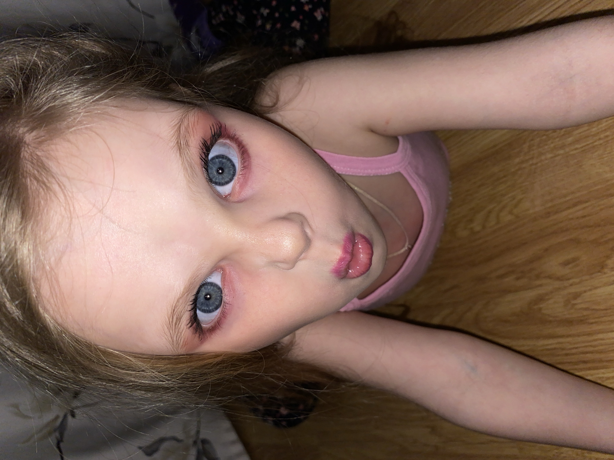 A fair-skinned girl with blonde hair and blue eyes is lying on her back with her head tilted towards the camera. She has pink eye makeup and lipstick applied. She is wearing a light pink tank top. The background shows a wooden floor, bedsheets with a floral pattern, and a piece of clothing with a dark floral pattern. There is no information to suggest that the image was taken in Votkinsk, Russia.