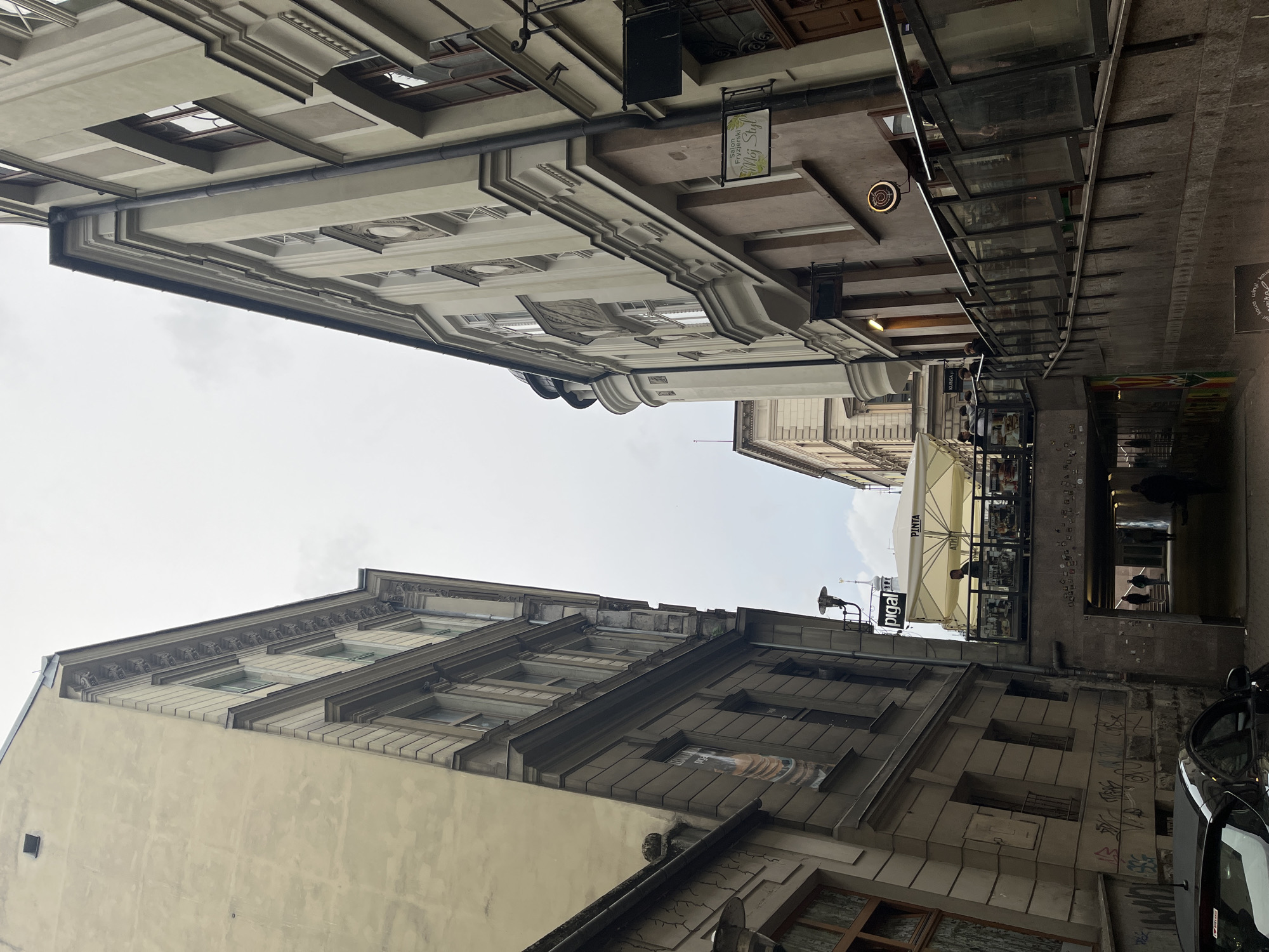 The image depicts a narrow street in an urban environment. Buildings with multi-story facades and architectural details such as cornices and windows flank both sides. The sky is visible between the buildings. A restaurant or cafe with outdoor seating under a canopy is present. An underpass is also visible, with people walking through it. A car is parked on the right side of the street. The scene suggests an older European city setting.