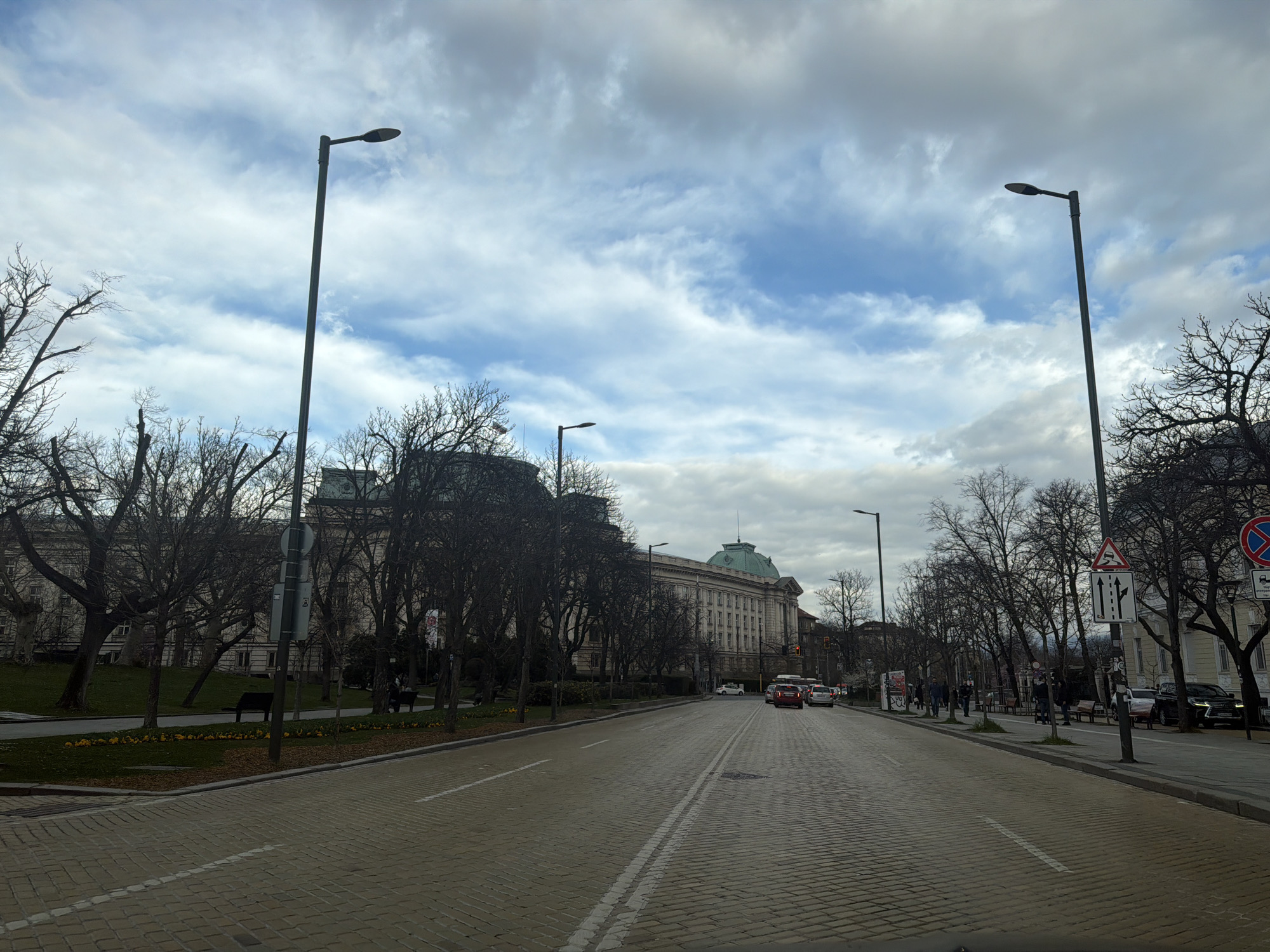 The image depicts a street in Sofia, Bulgaria, leading towards the National Assembly building, distinguishable by its green dome. The street is paved with cobblestones and marked with double white lines. Tall, bare trees line both sides of the street, interspersed with streetlights. A sidewalk runs alongside the road, with benches and a few people walking. Cars are visible on the road. Traffic signs are present on the right side of the street. The sky is overcast with a mix of grey and blue clouds.