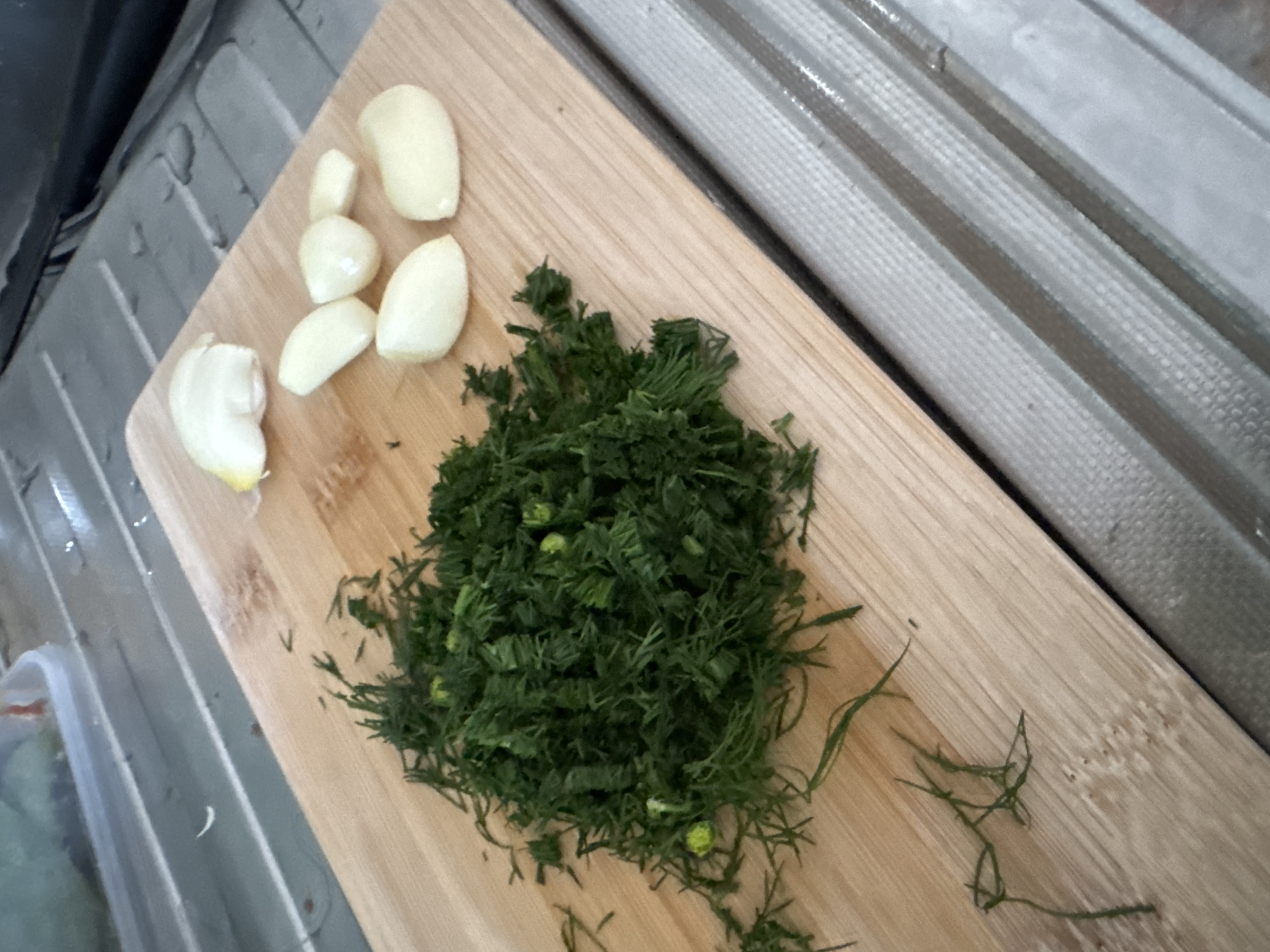 A wooden cutting board is placed on a metal surface with parallel ridges. On the cutting board, a pile of chopped fresh dill is to the left, and several peeled garlic cloves are to the right. In the upper left corner, a portion of a clear plastic container with a green substance is visible. The lighting suggests an indoor environment. No specific location in Höhenkirchen-Siegertsbrunn, Germany can be determined from the image.