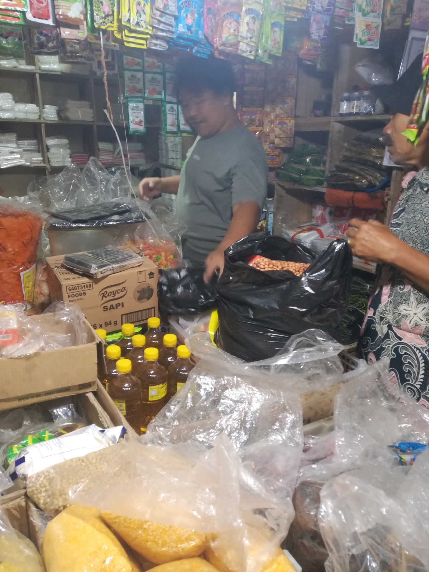 A medium shot captures a dimly lit indoor market stall in Garut, Indonesia. The foreground is cluttered with plastic-wrapped goods, including bags of yellow legumes and what appears to be cooking oil in numerous yellow-capped bottles. A cardboard box labeled "Royco SAPI" is prominently displayed, along with a calculator placed on top of it. In the mid-ground, a man with dark, curly hair and wearing a grey t-shirt is partially visible, looking to his right. To the right of the frame, a woman with dark hair and wearing a patterned garment is interacting with a large black plastic bag filled with peanuts. Behind the people, shelves are stocked with various packaged food items, including brightly colored snacks and sachets. The overall impression is one of a busy, perhaps somewhat disorganized, local market.
