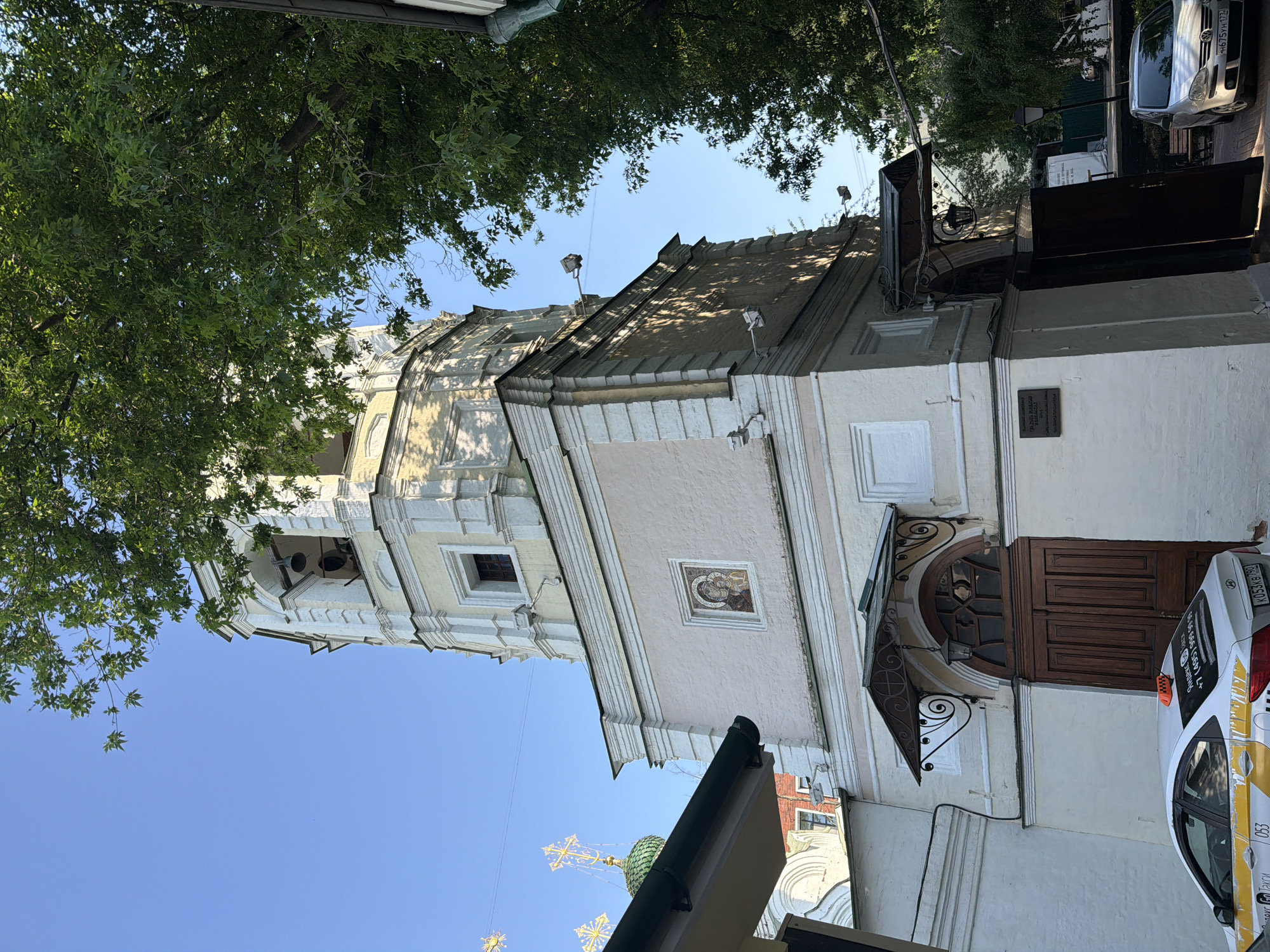The image shows a street-level view of the Church of St. Nicholas on Peski in Moscow, Russia. The architecture is a light-colored baroque style. A tall bell tower is visible on the left. The entrance to the church is on the right with a decorative overhang. A taxi is parked next to the church entrance. Green trees overhang the building at the top of the image.