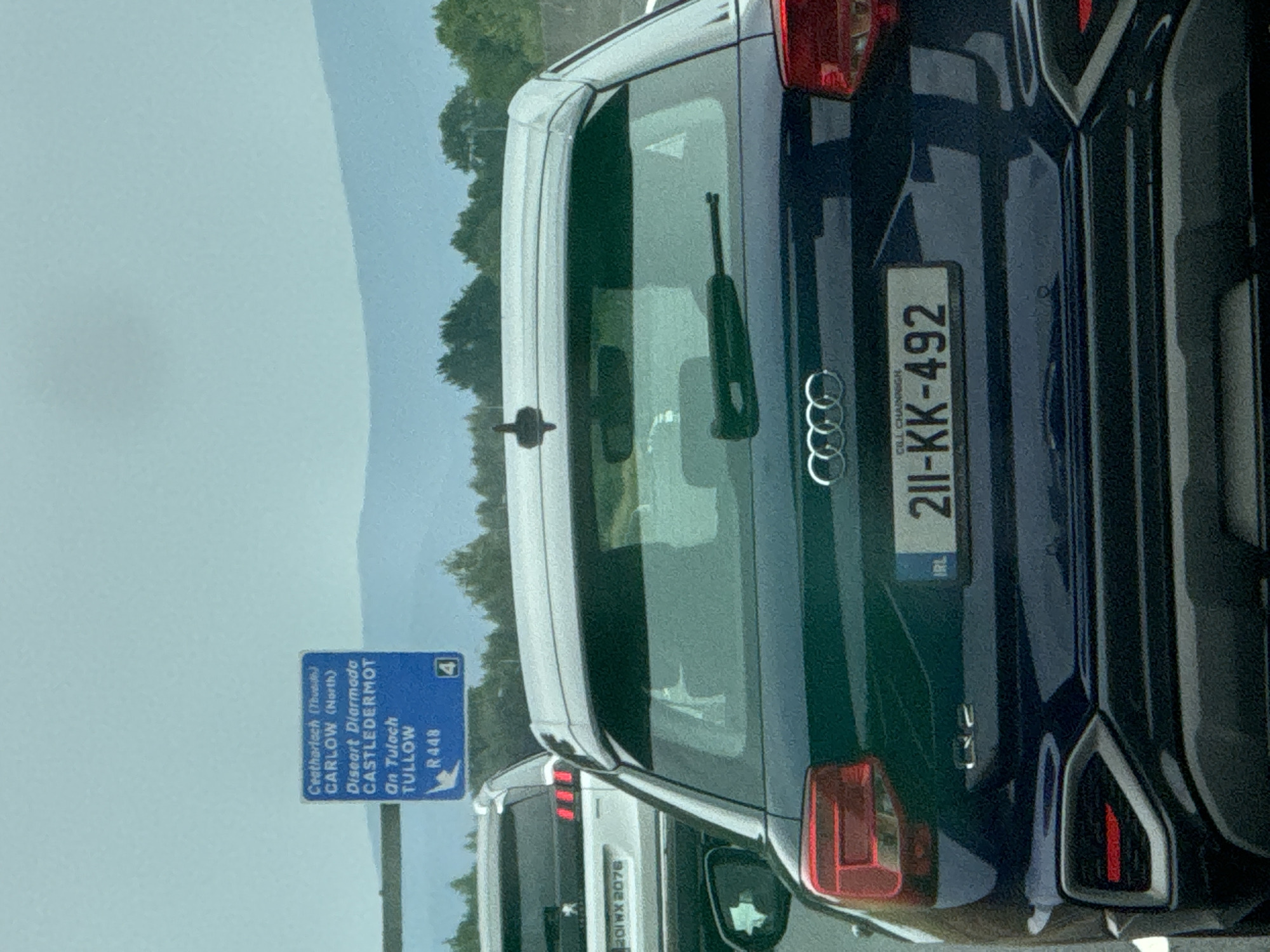 In the foreground, the rear of a dark blue Audi Q2 is visible, facing away from the camera. The license plate reads "IRL 211-KK-492". Behind the Audi, the rear of a white Peugeot SUV is visible, with a license plate that reads "201WX 2076". Further in the background, a blue road sign with white text indicates directions to "Ceatharlach (Thurles)", "CARLOW (North)", "Deseart Diarmada", "CASTLEDERMOT", "An Tuloch", "TULLOW", and "R448", with a directional arrow and a square symbol containing the number "4". The scene appears to be on a highway with green trees and rolling hills in the distance under a light blue sky. The image does not contain any people. The specific location of Pill Belan, Ireland is not indicated in the image.