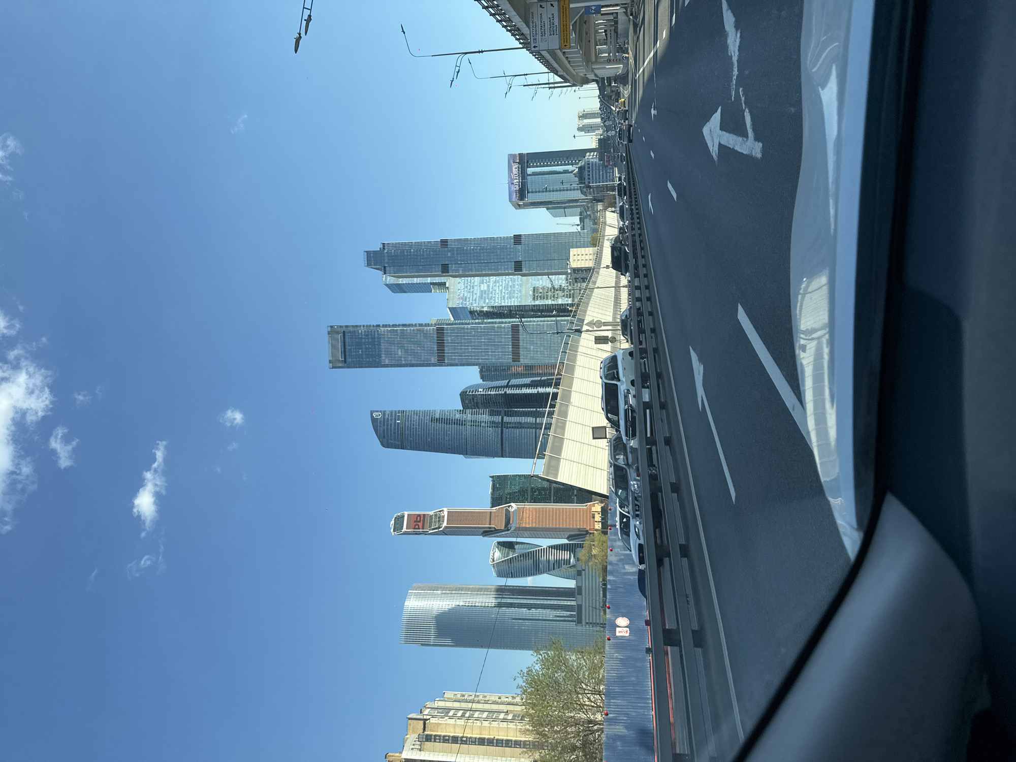 The image shows a view of Moscow City, a modern business district in Moscow, Russia, taken from inside a car on a road. Several skyscrapers of varying designs and heights are visible against a clear blue sky with a few scattered clouds. The road has multiple lanes and white lane markings, including an arrow indicating a direction change. There are other vehicles on the road, primarily white cars. A bridge or overpass is visible above the road. The interior of the car, including part of the dashboard and a window frame, is visible in the foreground.