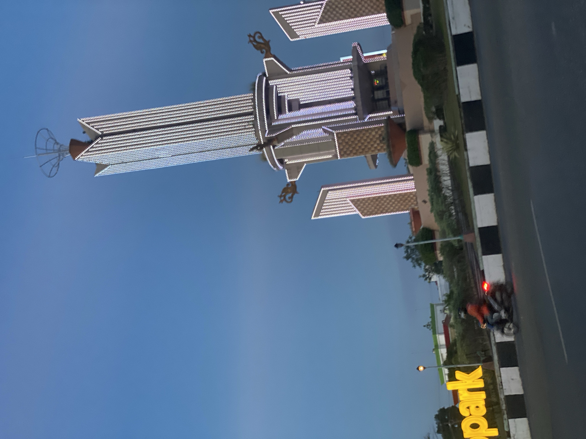 The image depicts the Tugu Lambang Sari monument in Banjarbaru, Indonesia, at dusk. The monument, illuminated by numerous white lights, is the central subject, rising prominently against a clear blue sky. Flanking the main tower are two smaller, similarly lit structures. A motorcycle with a rider wearing an orange jacket is seen in the foreground, moving from left to right. To the left of the motorcycle, a large yellow sign displaying the word "park" is visible. The scene is set on a paved road with a median strip marked with black and white stripes. Trees and other vegetation are present around the monument and along the roadside, suggesting a park-like setting. The lighting suggests it is either dawn or dusk.