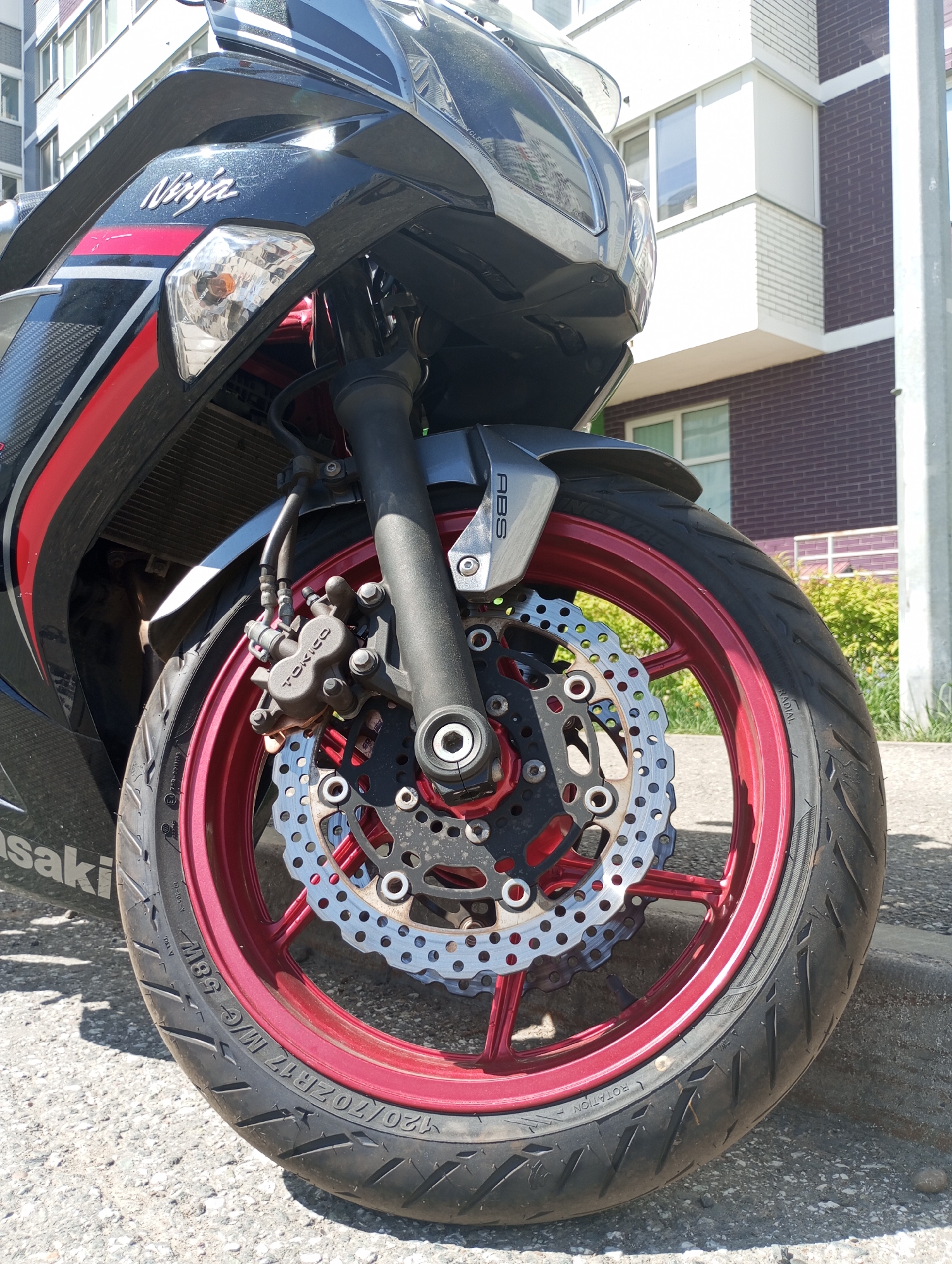 Here's a detailed description of the image:
**Subject:** A close-up view of the front wheel of a "Ninja" brand motorcycle. The wheel has a red rim and a black hub. A silver brake disc is visible with multiple holes. The tire is black and shows the markings "120/70ZR17 M/C 58W" along with "RADIAL" and "ROTATION". The brake caliper is labeled "TOKICO." The motorcycle has an ABS (anti-lock braking system) module above the wheel.
**Setting:** The motorcycle is parked on an asphalt surface next to a curb. Behind the motorcycle, a building is visible, which is light colored on its top level and has red brick on the ground level. There is some greenery in the background, suggesting a park or yard.
**Details:** The motorcycle appears to be in good condition. The photograph is taken in daylight.