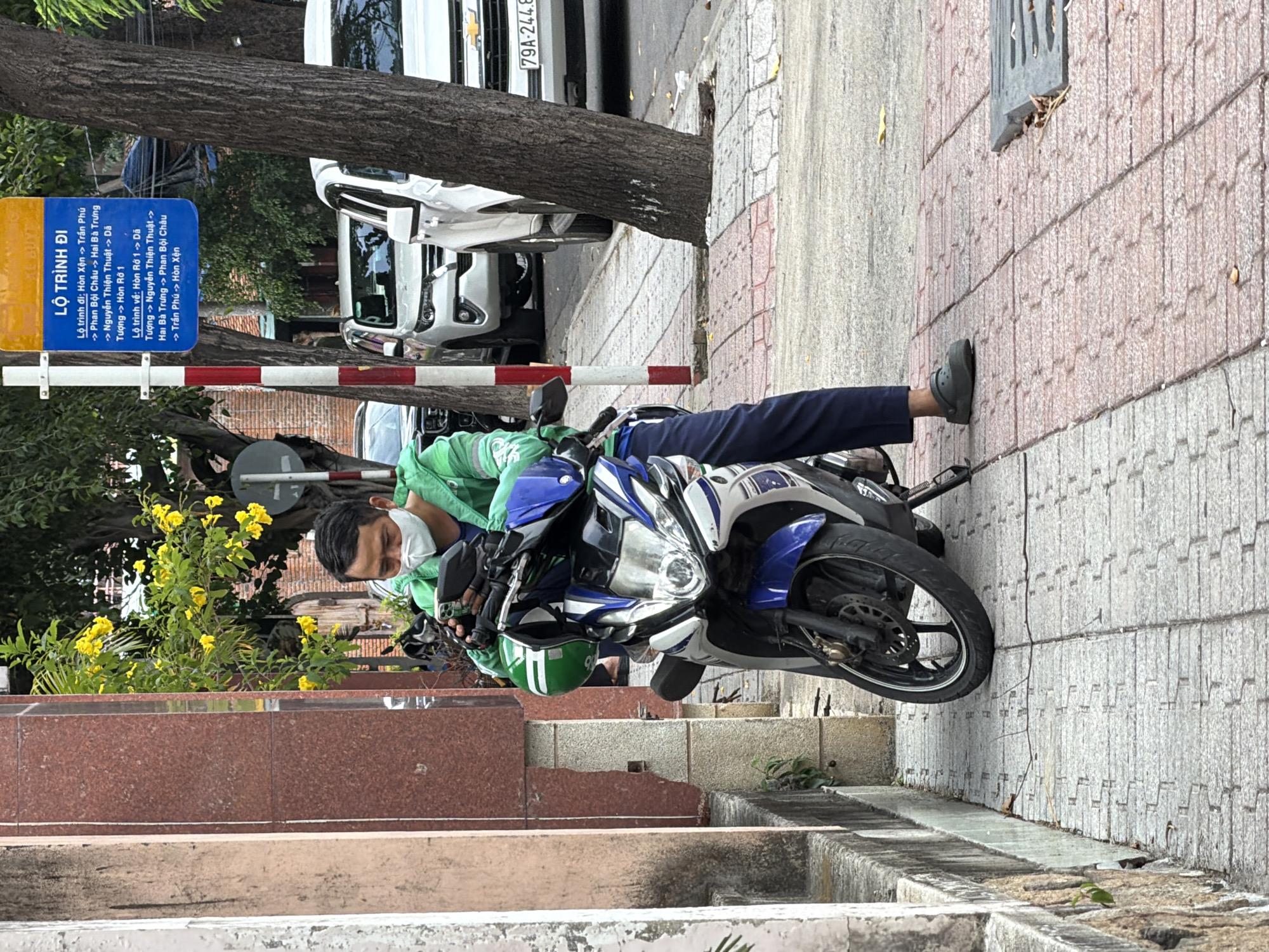 A man wearing a white mask and a green jacket is sitting on a blue and white motorcycle, looking at his phone. The motorcycle is parked on a sidewalk in Nha Trang, Vietnam. The sidewalk is paved with interlocking bricks and has a stone border. In the background, there are trees, cars, and a bus stop sign with Vietnamese text. The scene appears to be a typical urban street in Vietnam.