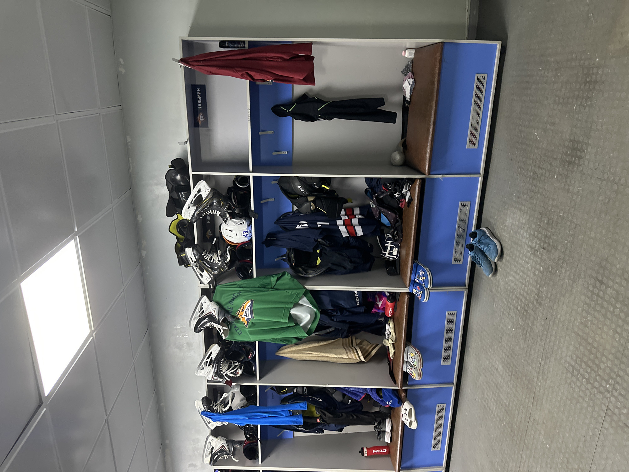 This image depicts an empty ice hockey locker room, viewed from a slightly elevated perspective. The central focus is a long row of blue and grey cubbies or stalls, each equipped with hooks for hanging clothes and a brown cushioned bench at the bottom.
No people are visible, but the stalls are filled with a variety of ice hockey equipment and personal belongings, suggesting recent use or preparation for a game or practice. Items include multiple pairs of ice hockey skates (many bearing the "BAUER" brand), several helmets, pads, and various articles of clothing such as jackets, jerseys (one green with an animal logo and some unreadable text), t-shirts, and towels. Water bottles, some branded "CCM" and "FLEX," are placed on shelves or benches. Several pairs of casual shoes, including sneakers and sandals, are scattered on the floor in front of the stalls.
The floor is a grey, textured surface, possibly rubber or composite, typical of sports facilities. The walls are light grey, and the ceiling features white square acoustic panels with integrated rectangular fluorescent light fixtures, providing bright, even illumination.
Visible text includes the name "КАЗЬМИН" (KAZ'MIN) on a dark plaque above one of the stalls, likely indicating a player's designated spot. The brand "CCM" is seen on a red water bottle, and "BAUER" on skates. The locker room appears functional and well-maintained, but the scattered equipment hints at the dynamic environment of a sports team. The time of day and weather are not discernible due to the indoor setting.