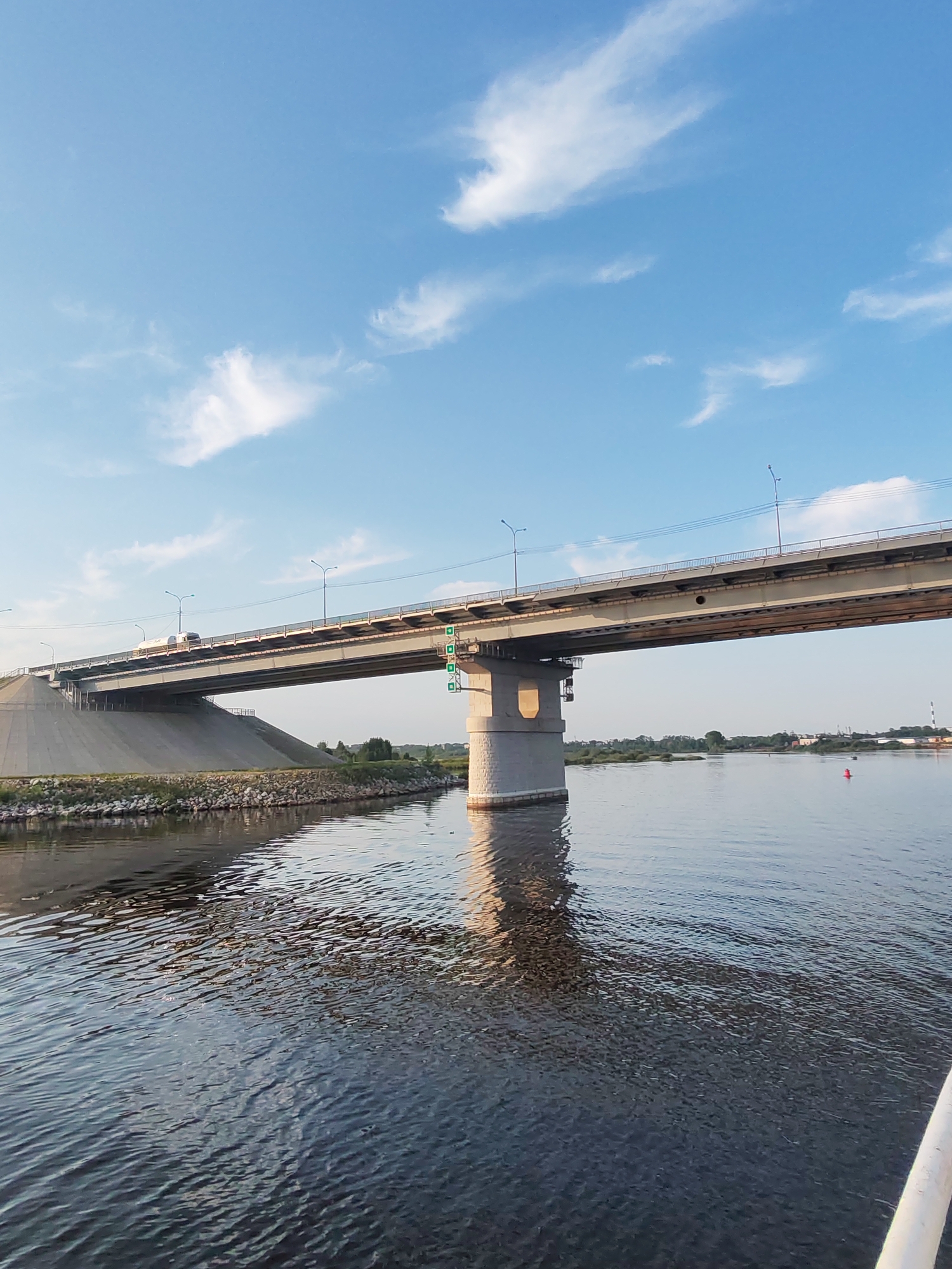 A wide shot captures a modern concrete and steel bridge spanning across a wide, calm river under a clear blue sky with scattered white clouds. The bridge's roadway is visible, with a vehicle, possibly a truck or large van, driving on it. The bridge's support structure, a large concrete pillar, is prominent in the foreground on the right side of the image. The riverbanks are lined with rocks and vegetation. On the far bank, a line of trees and some distant buildings are visible. A red buoy floats in the water to the right of the bridge. The image appears to be taken from a boat due to the perspective of the rippling water. The overall scene is one of a functional, modern infrastructure element integrated into a natural landscape. The location is identified as Sheksna, Russia.