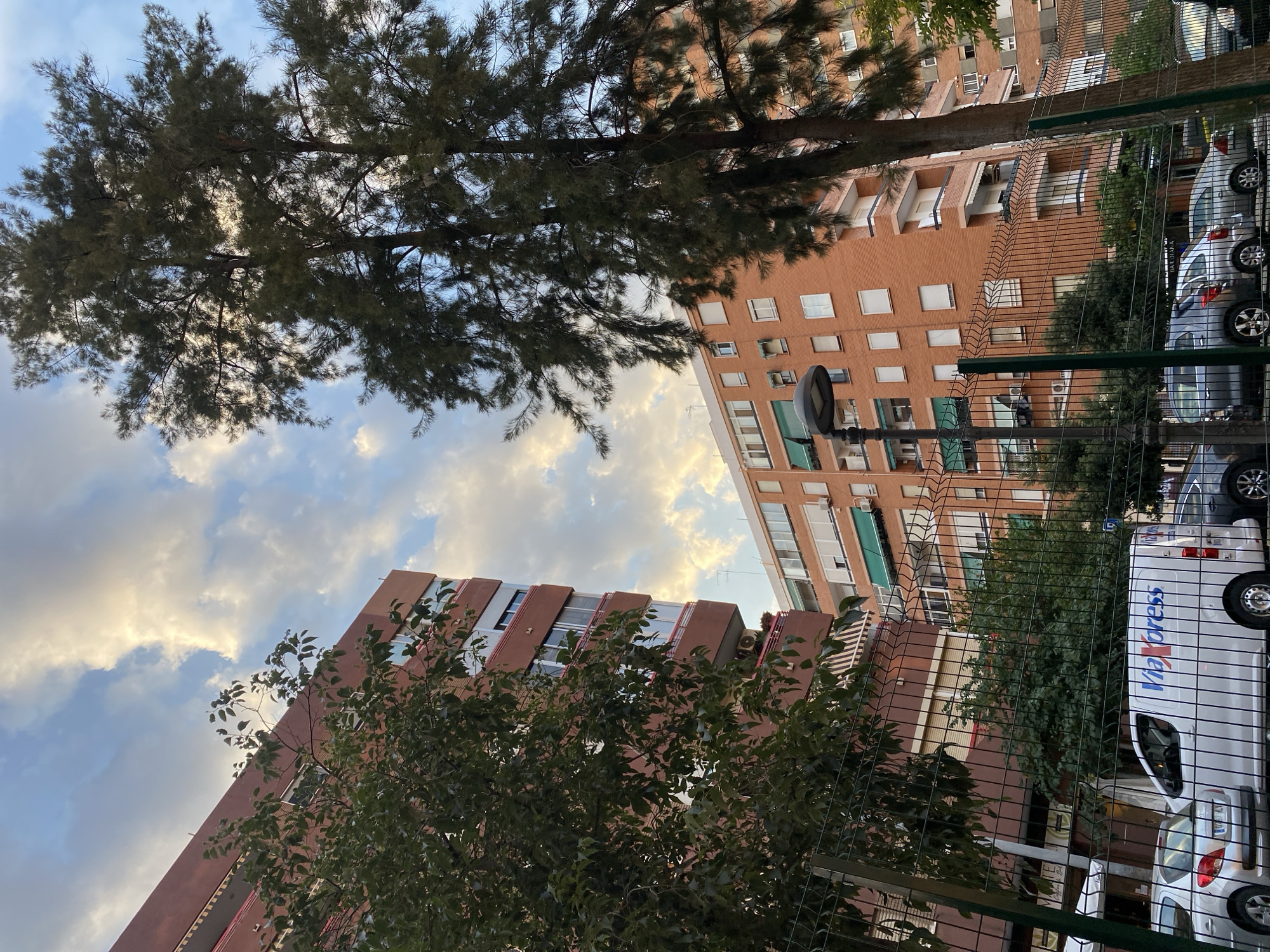This media file captures an outdoor scene in València, Spain, likely during the late afternoon or early evening as indicated by the warm, diffused sunlight filtering through the clouds and tree branches. The perspective is looking upwards, with two multi-story brick buildings dominating the frame.
The building on the left is a reddish-brown brick structure with multiple floors, featuring a pattern of balconies with red railings. The building on the right is also brick, a lighter shade of orange, with numerous windows, some of which have green awnings. Trees with dense green foliage are prominent in the foreground and midground, partially obscuring the buildings and creating a dappled effect. The sky above is a mix of blue and white clouds, with patches of sunlight suggesting a partly cloudy sky.
In the lower right portion of the image, a wire fence runs across, behind which several vehicles are parked. One vehicle is a white van with blue text that appears to read "Via Express". A silver car is also visible. The setting appears to be a residential or urban area with some greenery and parking facilities. The overall mood is calm and perhaps slightly serene, with the natural elements of trees and sky contrasting with the urban architecture.