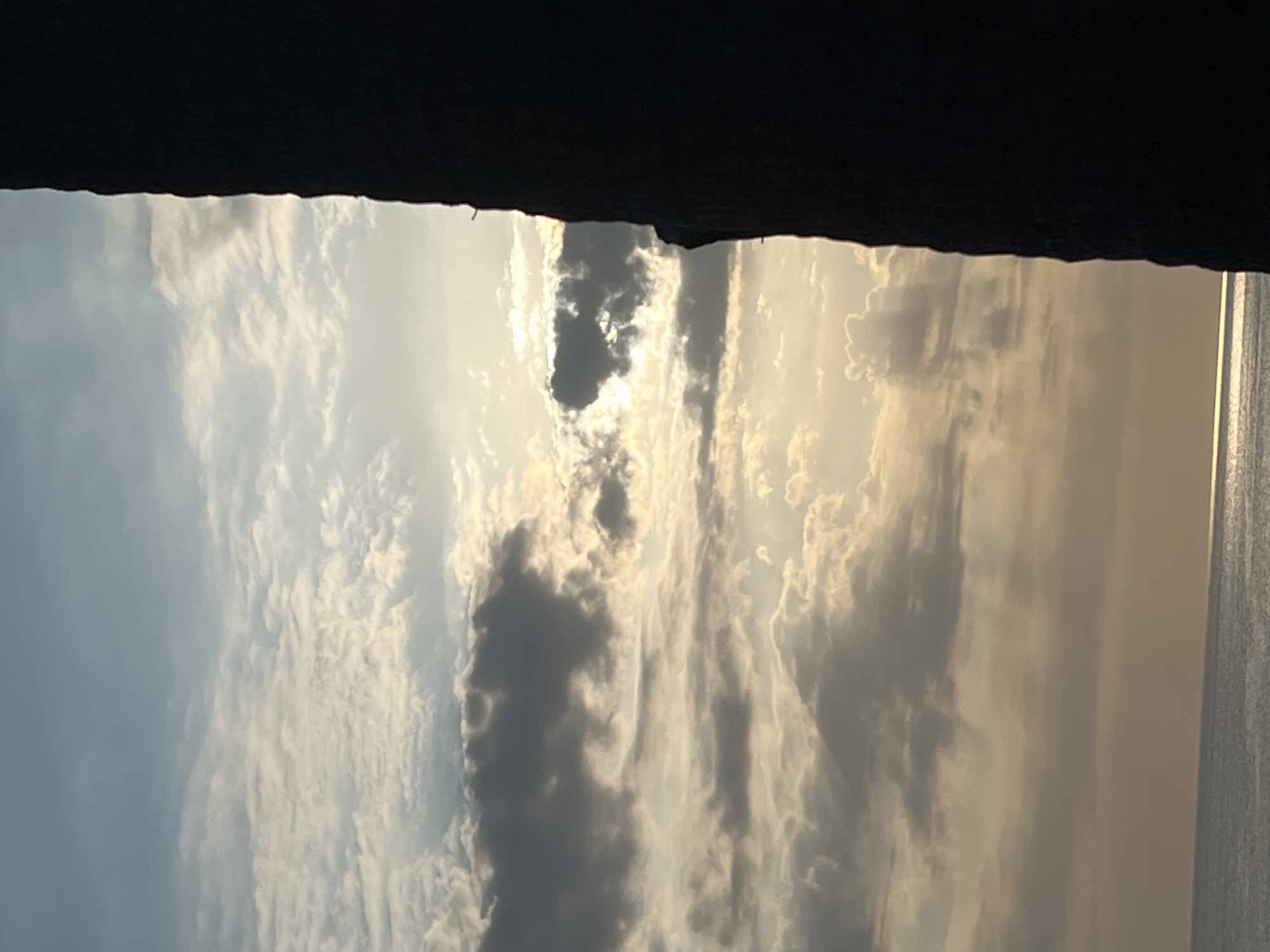 The image depicts a dramatic sky with scattered clouds at sunset, partially obscured by a dark, silhouetted structure on the right side. The sun is low on the horizon, casting a warm, golden light through the clouds, creating shafts of light and illuminating the water below. The sea is visible in the lower portion of the image, appearing calm with a shimmering reflection of the sky. The silhouetted structure, possibly a building or a natural formation, creates a strong vertical element that contrasts with the expansive sky and sea. The overall mood is serene and atmospheric, with a sense of natural beauty and dramatic lighting. This scene is characteristic of a sunset view from the coast of Kuta, Indonesia, a popular tourist destination known for its beaches and stunning sunsets.