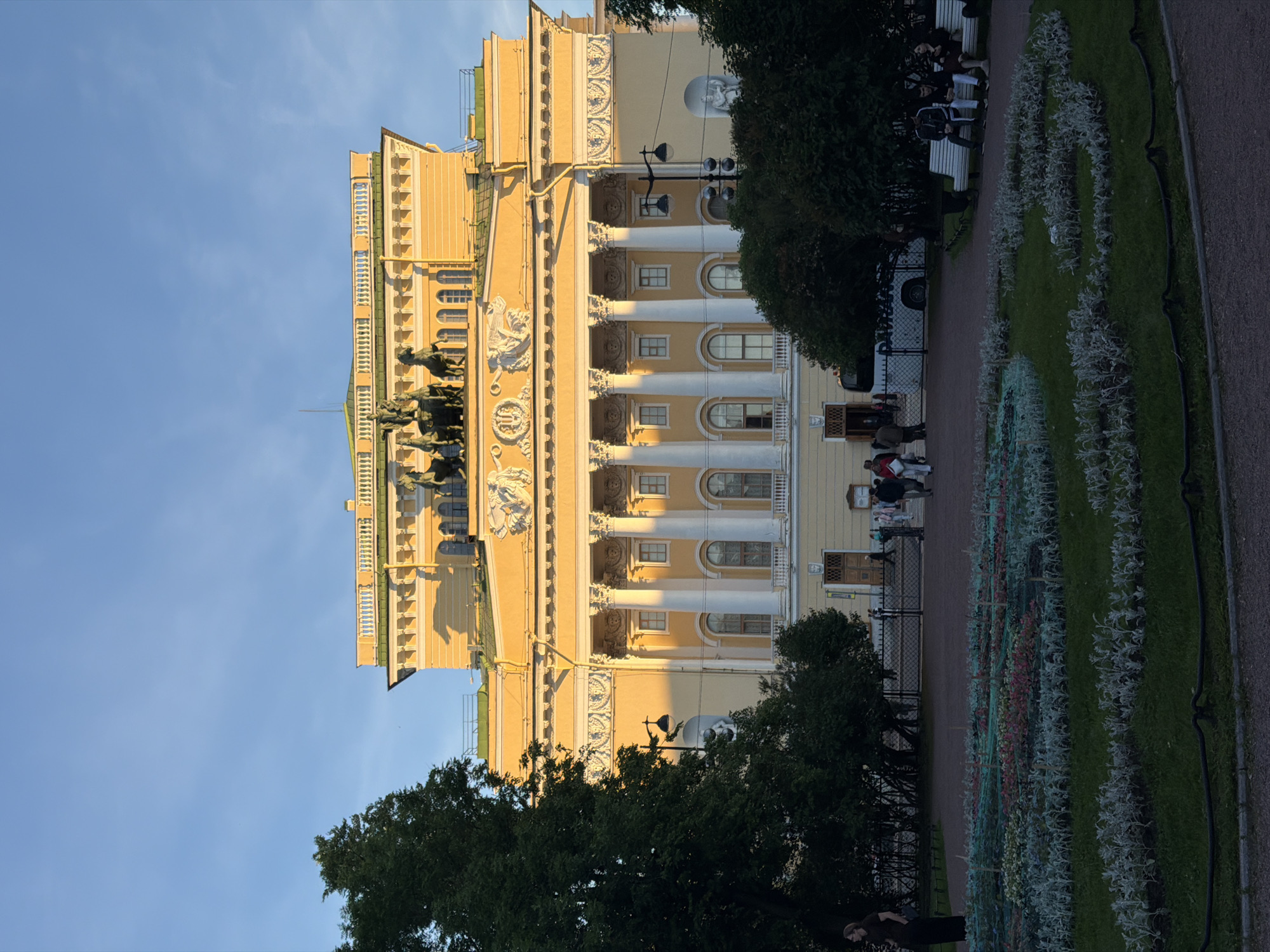 The image depicts the Alexandrinsky Theatre in St. Petersburg, Russia, bathed in the warm light of the late afternoon sun. The grand neoclassical building, painted a pale yellow, features a prominent portico with white Corinthian columns and a sculpted pediment. Above the entablature, a bronze quadriga, a chariot pulled by four horses, crowns the structure. Lush green trees flank the building, and in the foreground, a manicured garden with colorful flowerbeds and neatly trimmed grass is visible. Several individuals are present in the scene: a woman stands alone on the left, appearing to pose for a photo; a group of people are walking near the entrance of the theatre; and a few people are seated on a bench to the right, observing their surroundings. The sky above is a clear, vibrant blue, with a few wispy clouds. A white bus is parked near the theatre entrance, and a fence separates the garden from the paved walkway.