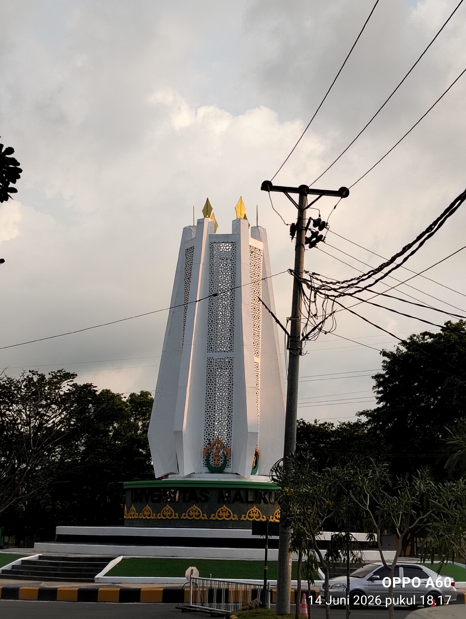 A wide-angle, low-light exterior shot captures the University of Malikussaleh monument in Reuleut Timu, Indonesia. The monument, a tall white structure with gold accents and intricate geometric cutouts, is the central focus. A utility pole with numerous wires runs diagonally across the upper right portion of the frame, obscuring parts of the monument and the sky. Dense green trees frame the base of the monument and extend into the background. The sky is overcast with diffused light. A silver sedan is parked near the monument on a paved road with yellow and black striped markings. The bottom right corner of the image contains a timestamp and device information: "OPPO A60 14 Juni 2026 pukul 18.17".