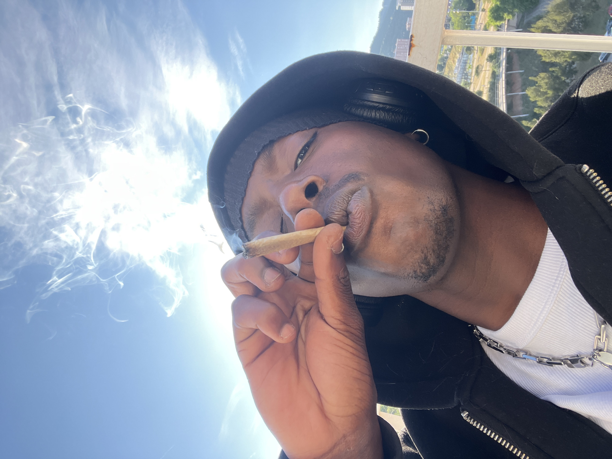 A man with dark skin is holding a lit cigarette in his right hand. He has short facial hair and is wearing a black hoodie over a white shirt. The man also wears a chain around his neck and headphones over his hood. A cigarette hangs between his lips and fingers, emitting a cloud of smoke. The photo is taken from a low angle, with a clear blue sky and wispy clouds in the background. In the distance, buildings and greenery suggest the location is in the city of Barcelona, Spain.