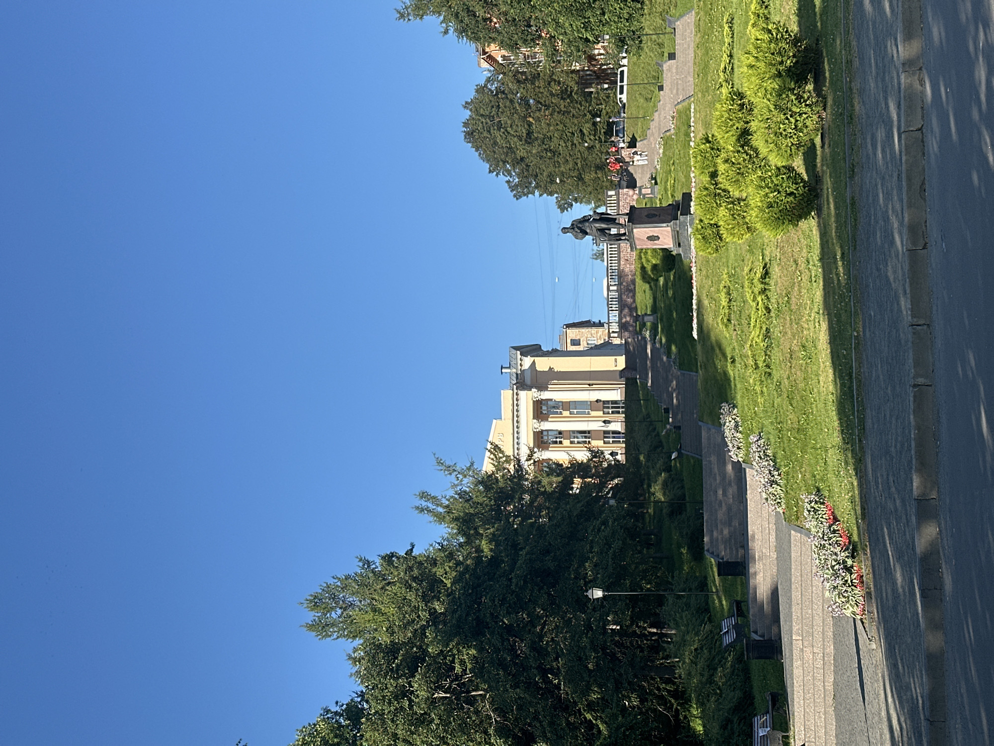 The image depicts a daytime view of a park in Petrozavodsk, Russia. In the foreground, a paved walkway is visible, with a curb separating it from a grassy area. Several manicured, spherical green bushes are arranged on the lawn. Behind the bushes, a wide, tiered stone staircase ascends a gentle slope. Along the sides of the staircase, flowerbeds with pink and red flowers add color to the scene. Benches are placed on landings along the stairs.
Further up the slope, a bronze statue of a man seated on a pedestal stands prominently. Beyond the statue, more stairs lead towards a light yellow building with classical architectural elements, including columns and large windows. This building appears to be a public institution. Dense green trees flank the stairs and the building. In the background, other buildings and more trees can be seen, under a clear blue sky. A few people are visible walking on the stairs and near the buildings. Street lamps are also present along the pathways. The overall impression is that of a well-maintained urban park with historical and architectural significance.
