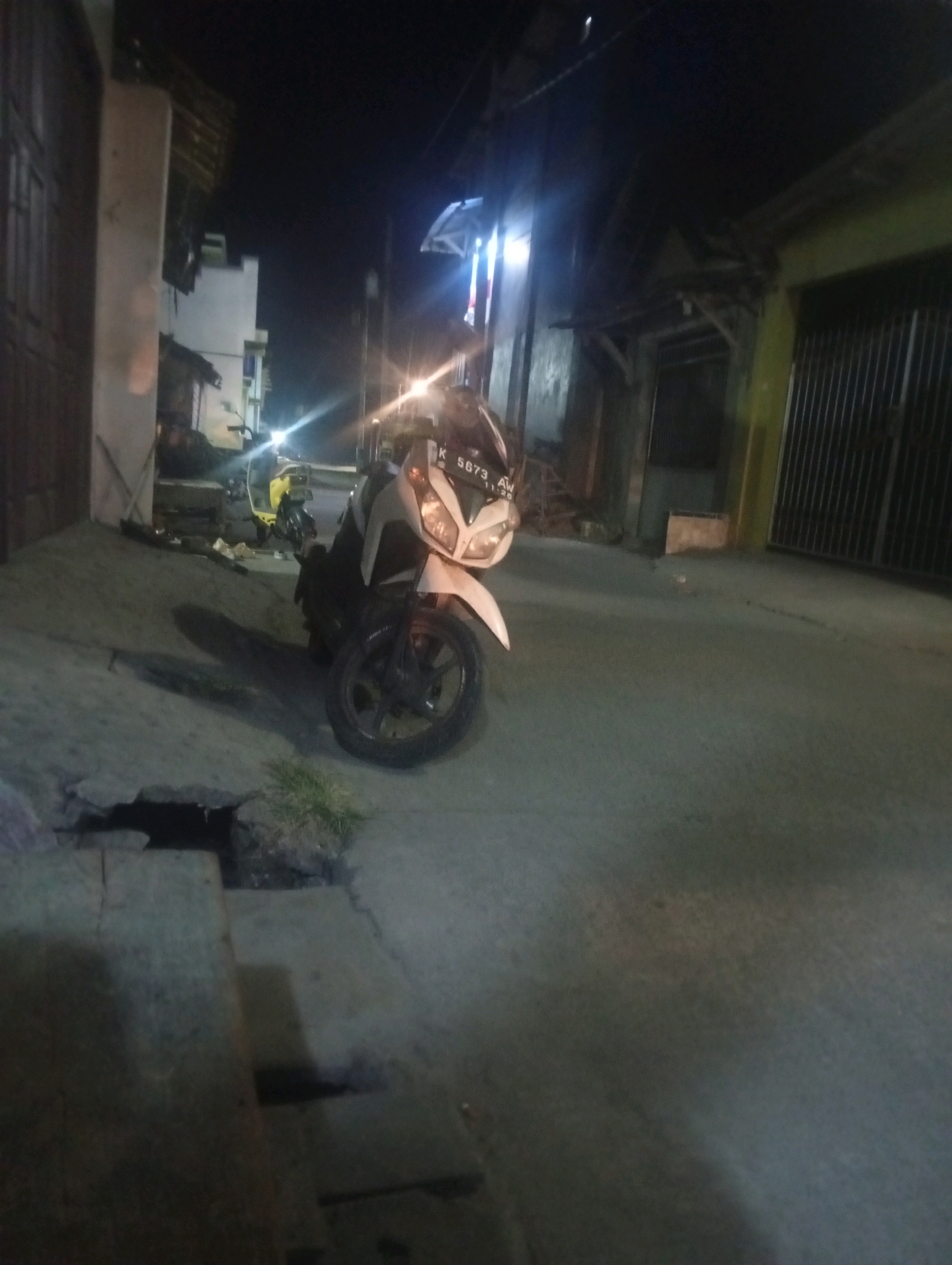 A scooter is parked on a street at night in Bajingjowo, Indonesia. The scooter is white and black, with a license plate that reads "K 5673 AW". It is positioned on the right side of the image, facing towards the viewer. The street is paved and appears to be an alleyway or side street, with buildings visible on either side. The lighting suggests it is nighttime, with some artificial lights illuminating the scene. There is a yellow scooter parked behind the white scooter. A dark opening in the pavement is visible in the lower left of the image.