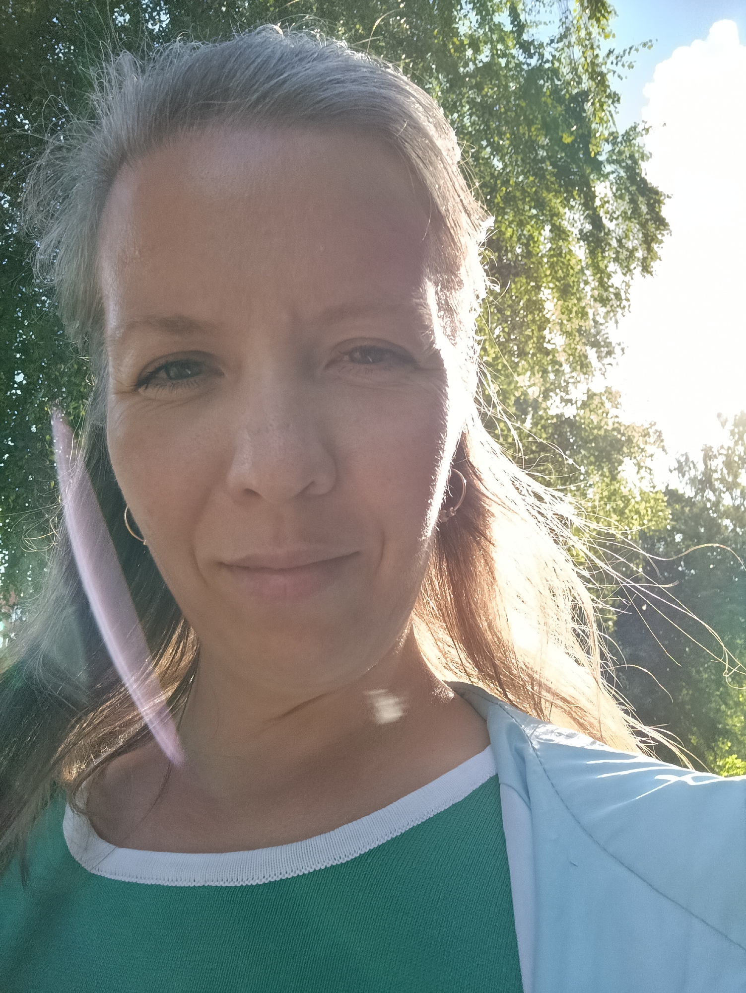 A selfie taken outdoors in daylight. The subject is a woman with fair skin, graying hair, and a slight smile. She is wearing a green shirt with a white trim and a light blue jacket. A light flare obscures part of her face and hair on the left side. The background is a green blur of trees and foliage, suggesting a park or natural setting. The location is not explicitly discernible as Chaikovsky, Russia from the image alone.