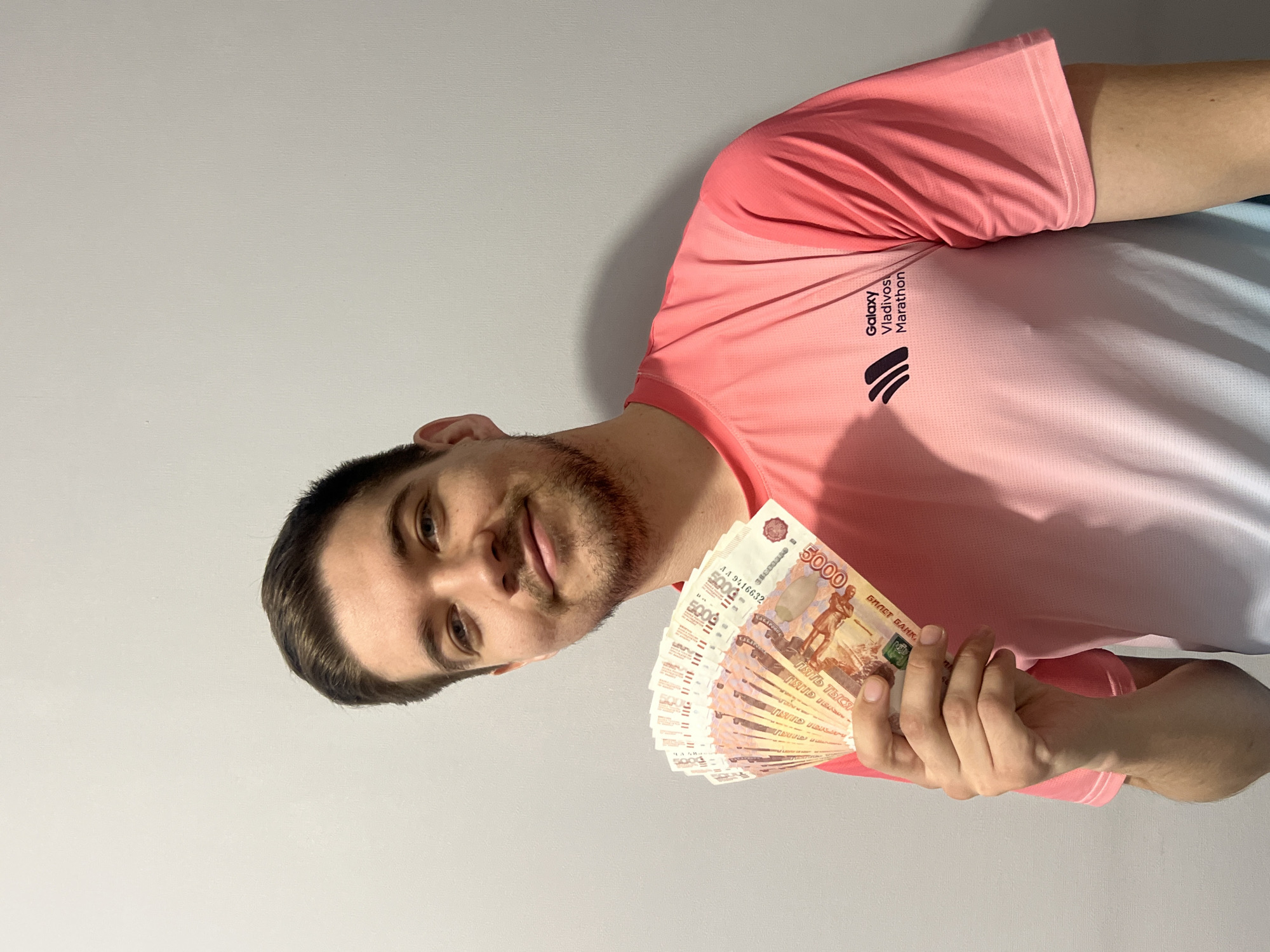A man is holding a stack of 5,000 ruble notes. He is wearing a pink and white Galaxy Vladivostok Marathon t-shirt. The background is a light grey or white wall. The man's location is uncertain.