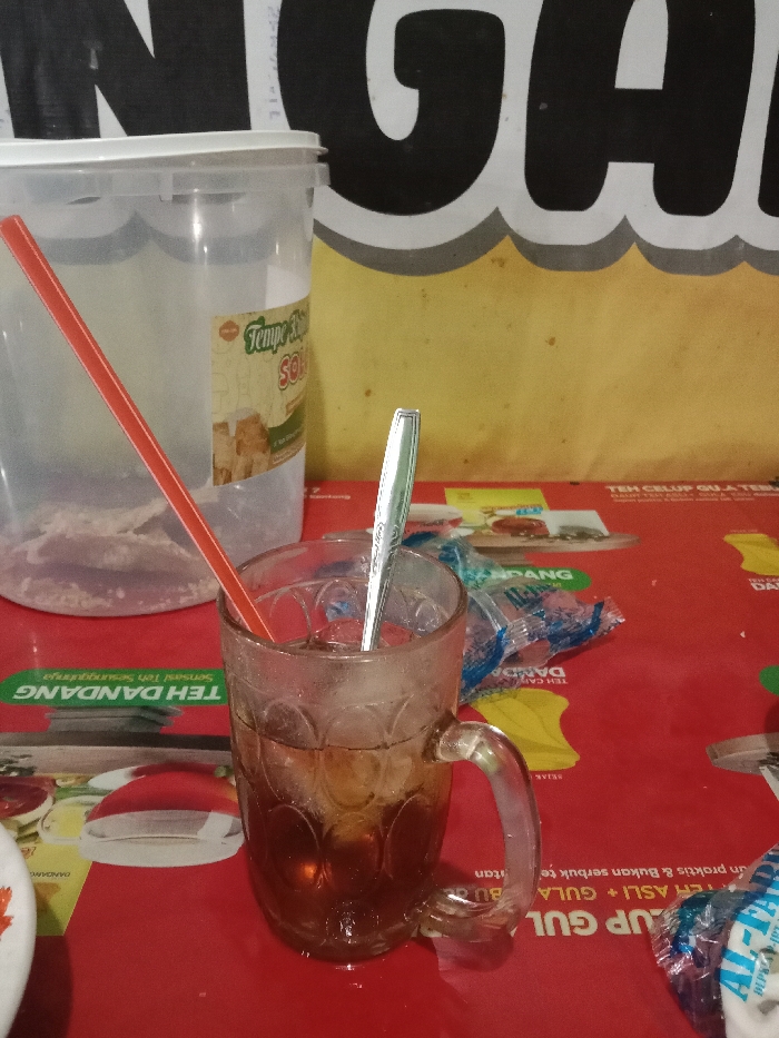 A glass of iced tea with a metal spoon inside sits on a red table in a small shop in Tugu, Indonesia (latitude: -6.975, longitude: 110.332). A red straw is in the glass, and a clear plastic container with a label that reads "Tempe & Soy Sauce" sits behind it. The table is covered in a red plastic tablecloth with a variety of product packaging, including a bag of sugar and a pack of "Teh Celup Gula" (tea with sugar). The wall behind the table is yellow and decorated with a large red-lettered sign that reads "ANG." The shop appears to be a small food stall or a warung.