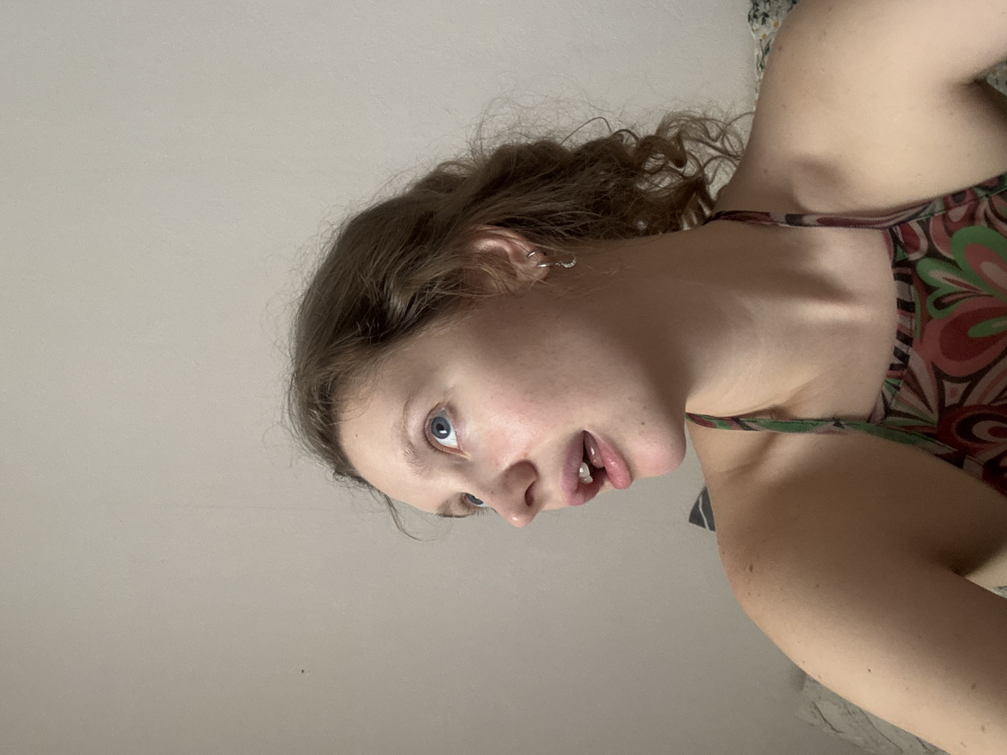 A young woman with light skin and curly brown hair is looking up and to the left. She is wearing a multi-colored halter top. Her mouth is slightly open, and she has a small earring in her left ear. The background is a plain, light-colored wall. There is no visible information to suggest the location is Izhevsk, Russia.