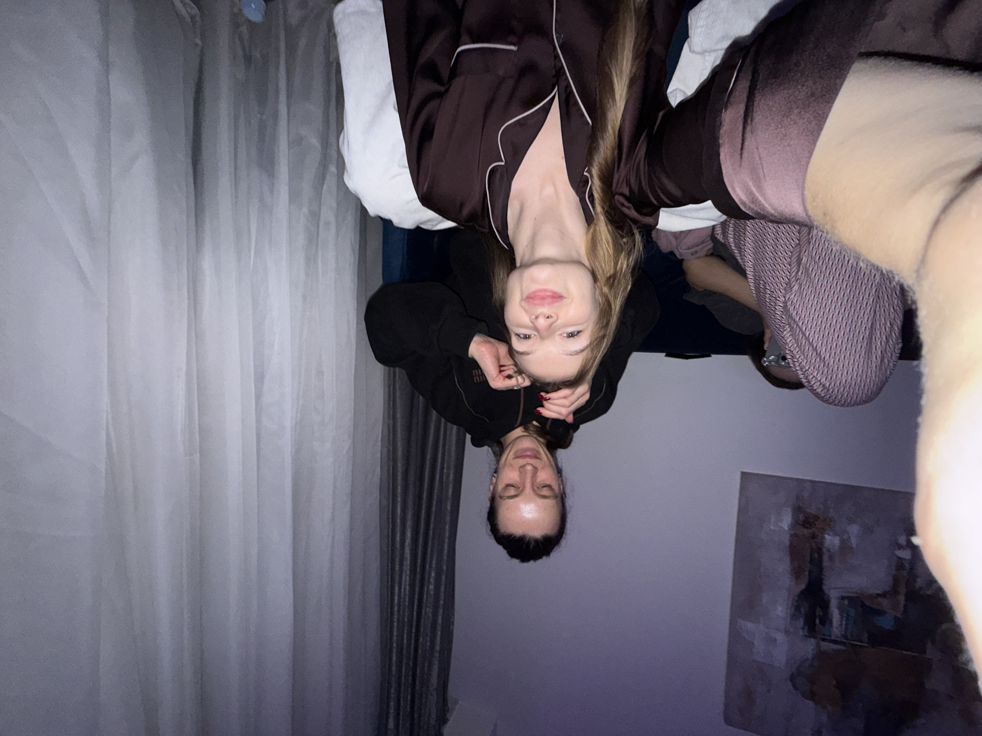 A woman in brown pajamas is lying in bed with a man. They are looking at the camera. The woman is holding the man's head. There is a painting on the wall and white curtains. The location is unknown.