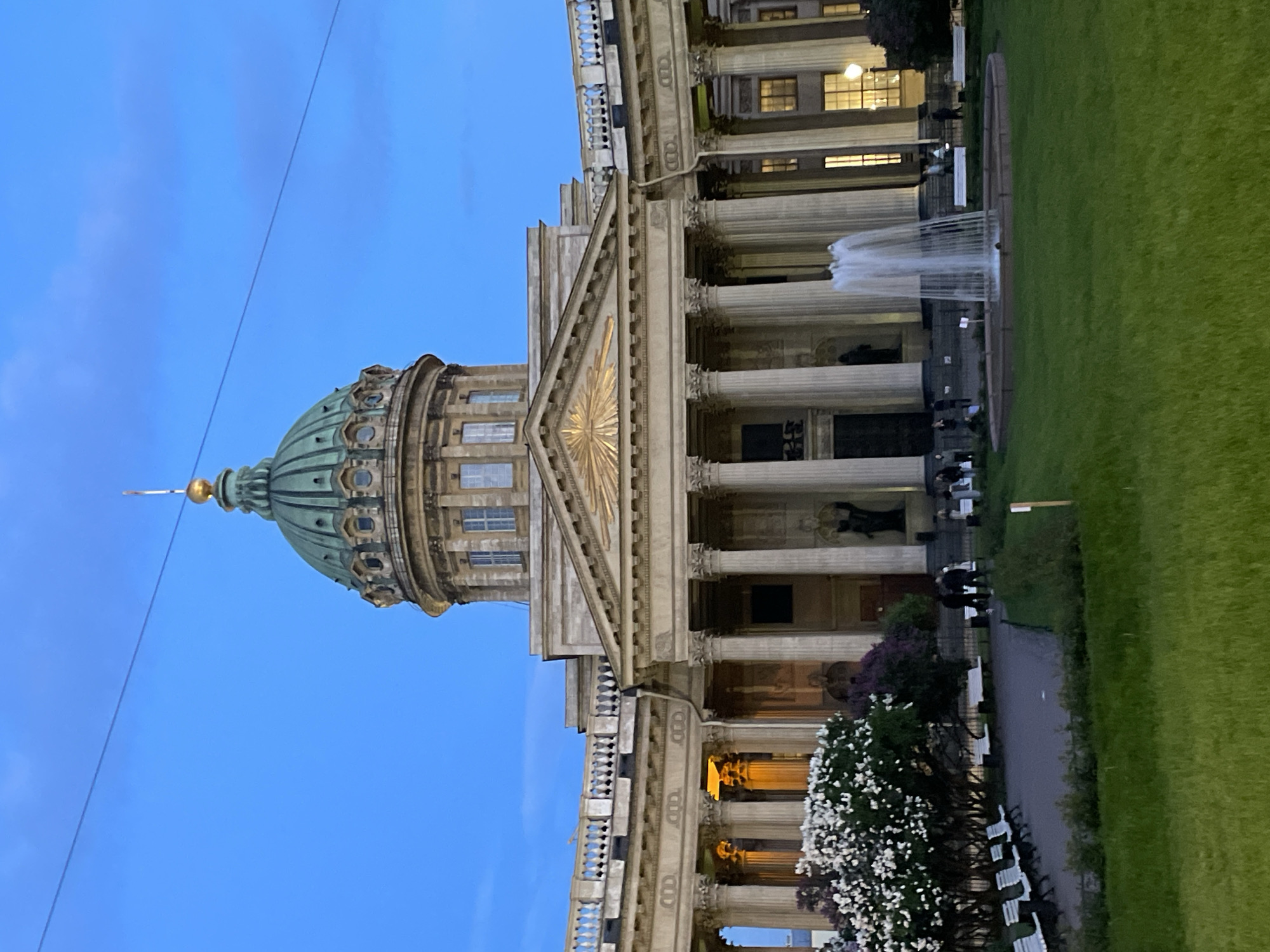 The image captures the majestic Kazan Cathedral in Saint Petersburg, Russia, at dusk or early evening. The grand neoclassical building dominates the left side of the frame, featuring a prominent, verdant green dome topped with a golden finial. Below the dome, a magnificent colonnade of tall, classical columns supports a triangular pediment, which is adorned with a striking golden sunburst motif at its center. Illumination from within the building and strategic lighting highlight architectural details, casting a warm glow on the portico and visible windows.
In front of the cathedral, a manicured green lawn stretches towards the right. A circular fountain, actively spraying water upwards, is centrally located on the lawn, drawing the eye. Beyond the fountain, a paved pathway meanders through the grass, lined with several white benches. Lush bushes and trees, some adorned with delicate white blossoms, frame the path and the base of the building. Scattered figures of people can be seen casually walking or standing in the area, enjoying the serene evening atmosphere.
The sky overhead is a clear, deep blue, transitioning from twilight to night, with no clouds visible. The overall scene conveys a sense of calm and grandeur, showcasing a significant historical and architectural landmark during a peaceful time of day. No discernible text is visible in the image.