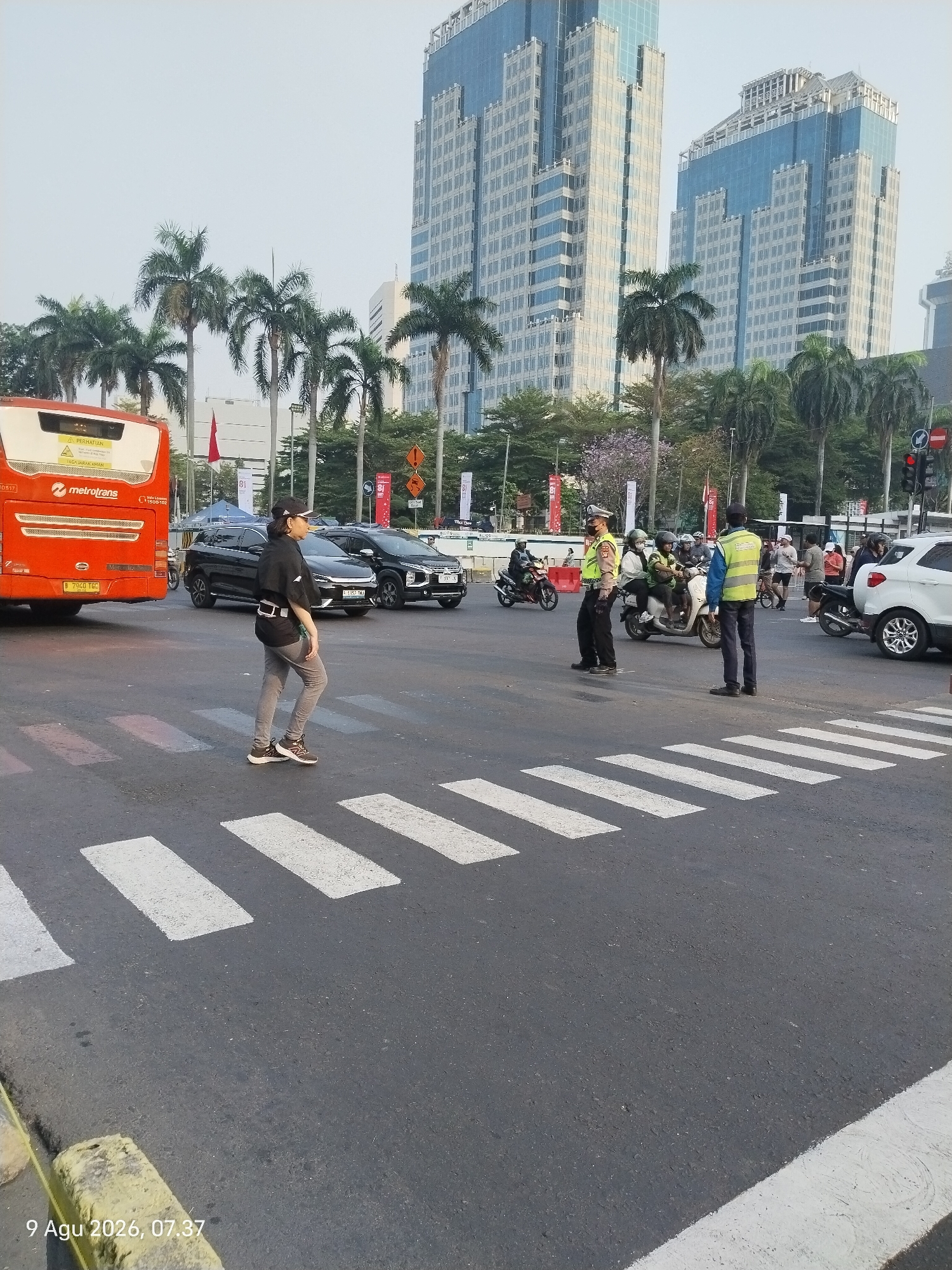 The image depicts an intersection in Central Jakarta, Indonesia, near high-rise buildings and palm trees. A red Metrotrans bus is parked on the left, facing away from the viewer. A pedestrian, wearing a black jacket and gray pants, is crossing a zebra crossing in the foreground. Two traffic police officers are visible, one in a dark uniform and the other in a yellow vest, directing traffic. Several vehicles, including cars and motorcycles, are present at the intersection, some of which are stopped. The sky is overcast. The timestamp "9 Agu 2026, 07.37" is visible in the lower left corner.