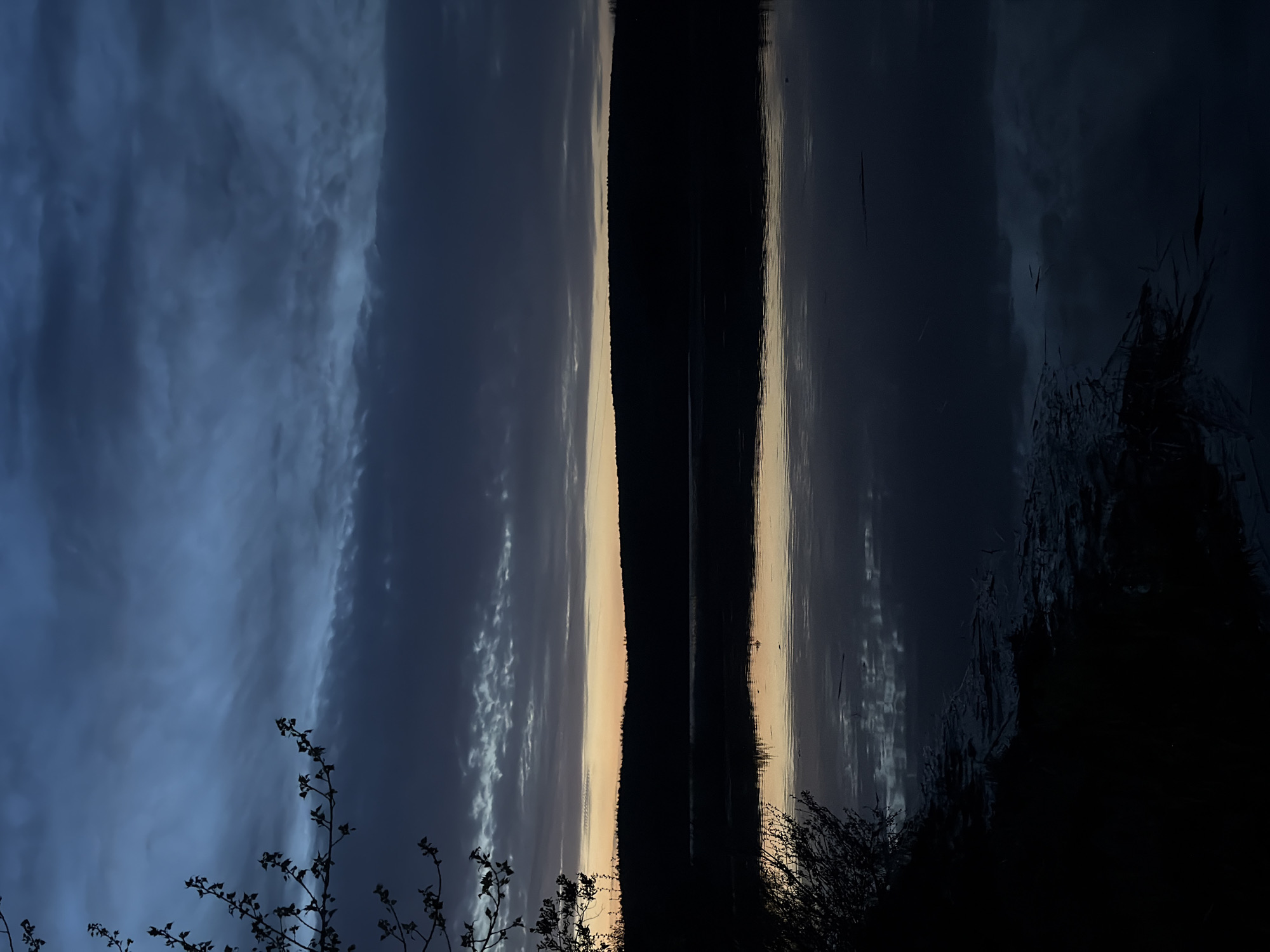 The image shows a nighttime landscape with a body of water reflecting the sky and a landmass. Dark clouds fill the sky on the left, transitioning to a sliver of lighter color along the horizon near the center of the image. This sliver is mirrored in the water. Silhouetted trees or bushes are visible in the foreground along the bottom of the frame. The scene is dark, with limited detail discernible in the shadows. The location is specified as Chaikovsky, Russia.