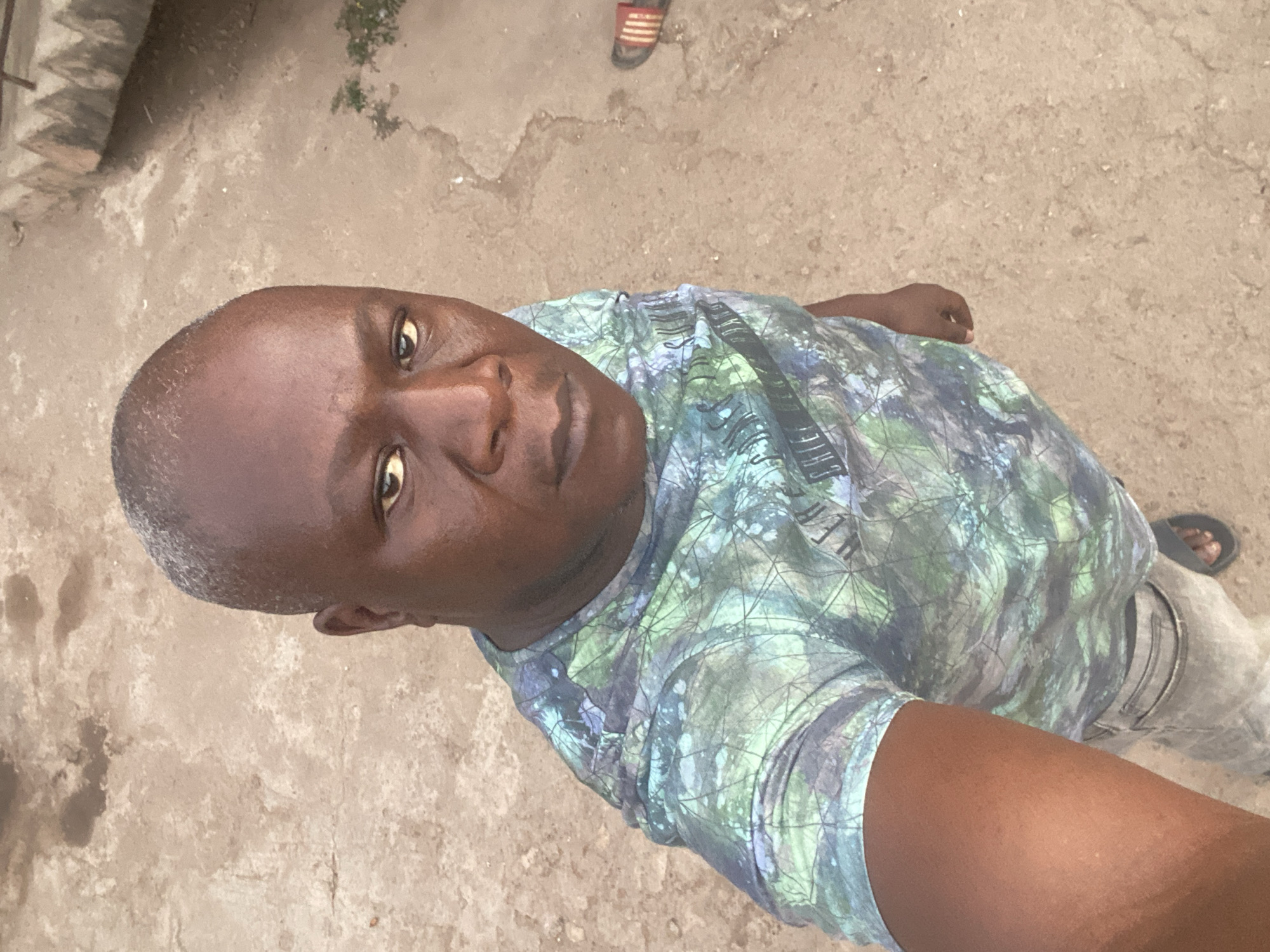 A man is standing outdoors, appearing to take a selfie. He has dark skin, a bald head with some graying hair, and is wearing a light blue and green patterned shirt. He is partially visible in the image, with part of his arm and leg also in the frame. The ground is a light gray concrete or stone surface. In the distance, a sandal is visible. The location is Sabon Tasha, Nigeria.