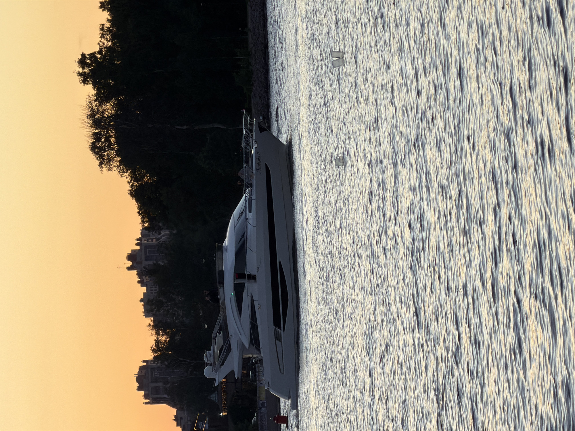 A large white yacht, named "Arabeska II", sails on a body of water during sunset. The sky is a warm orange hue, and the silhouette of trees and buildings lines the background. Two figures are visible on the yacht's deck. The water has a choppy surface, reflecting the colors of the sky. Two floating objects, possibly buoys or debris, are visible in the foreground. The scene likely depicts a view along the Neva River in Saint Petersburg, Russia, given the architectural style of the buildings in the background and the common sight of yachts on the river during warmer months.