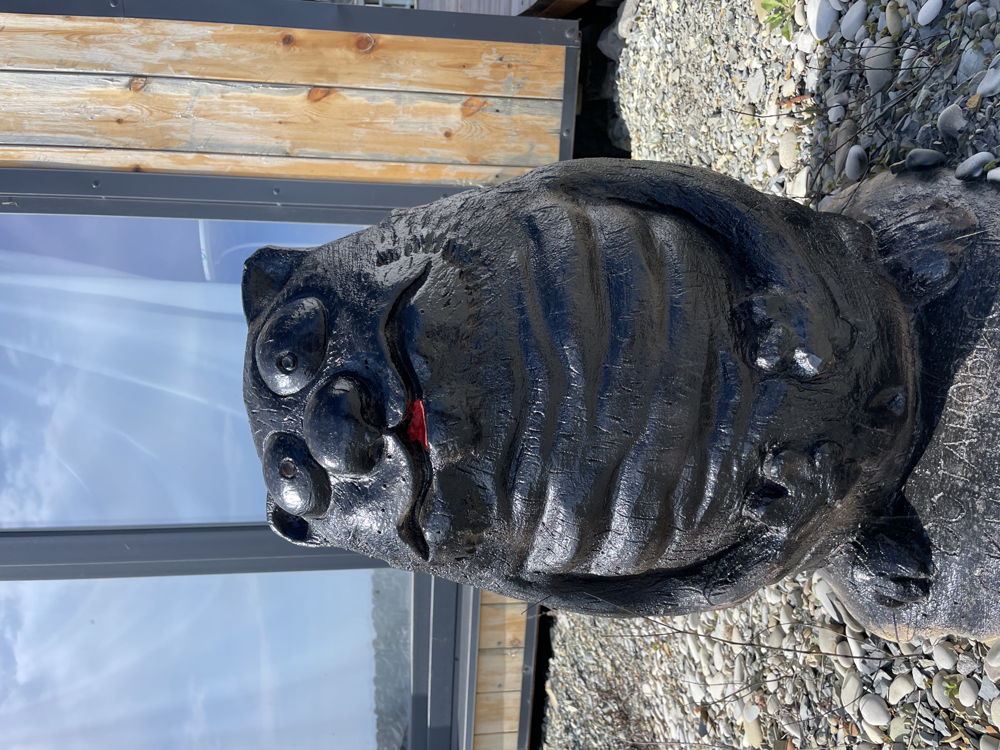 The image shows a black, glossy statue of a face, possibly a bear or feline, with red painted lips, placed on a bed of small white and grey stones. The statue is positioned outdoors, adjacent to a building with light wood paneling and a large reflective glass window showing a clear sky. The location is Olginka, Russia.