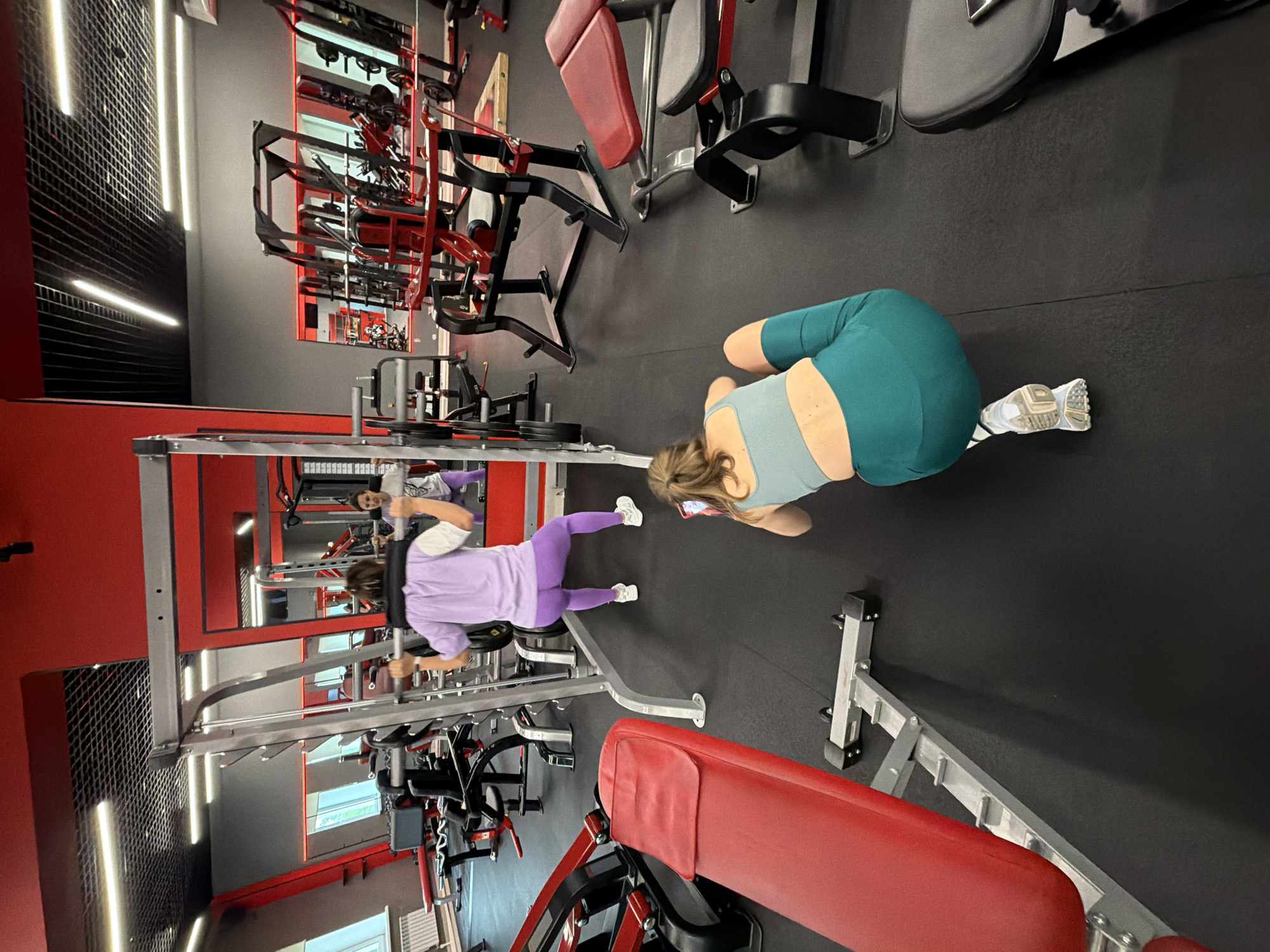 The image depicts the interior of a modern gym located in Birobidzhan, Russia. In the foreground, a woman is crouched on the floor, holding a smartphone and appearing to take a picture or record a video. She is wearing a teal sports bra and teal leggings. Behind her, another woman is performing squats with a barbell on her back, utilizing a squat rack. She is dressed in a lavender t-shirt and matching lavender leggings. Both women are positioned on a black rubberized floor, typical of gym environments. The gym features red and gray walls, a black ceiling with strip lighting, and various weightlifting equipment, including squat racks, benches, and weight machines, predominantly in red and black. Mirrors line one of the walls, reflecting the gym space and equipment. The overall impression is of a well-equipped fitness facility.