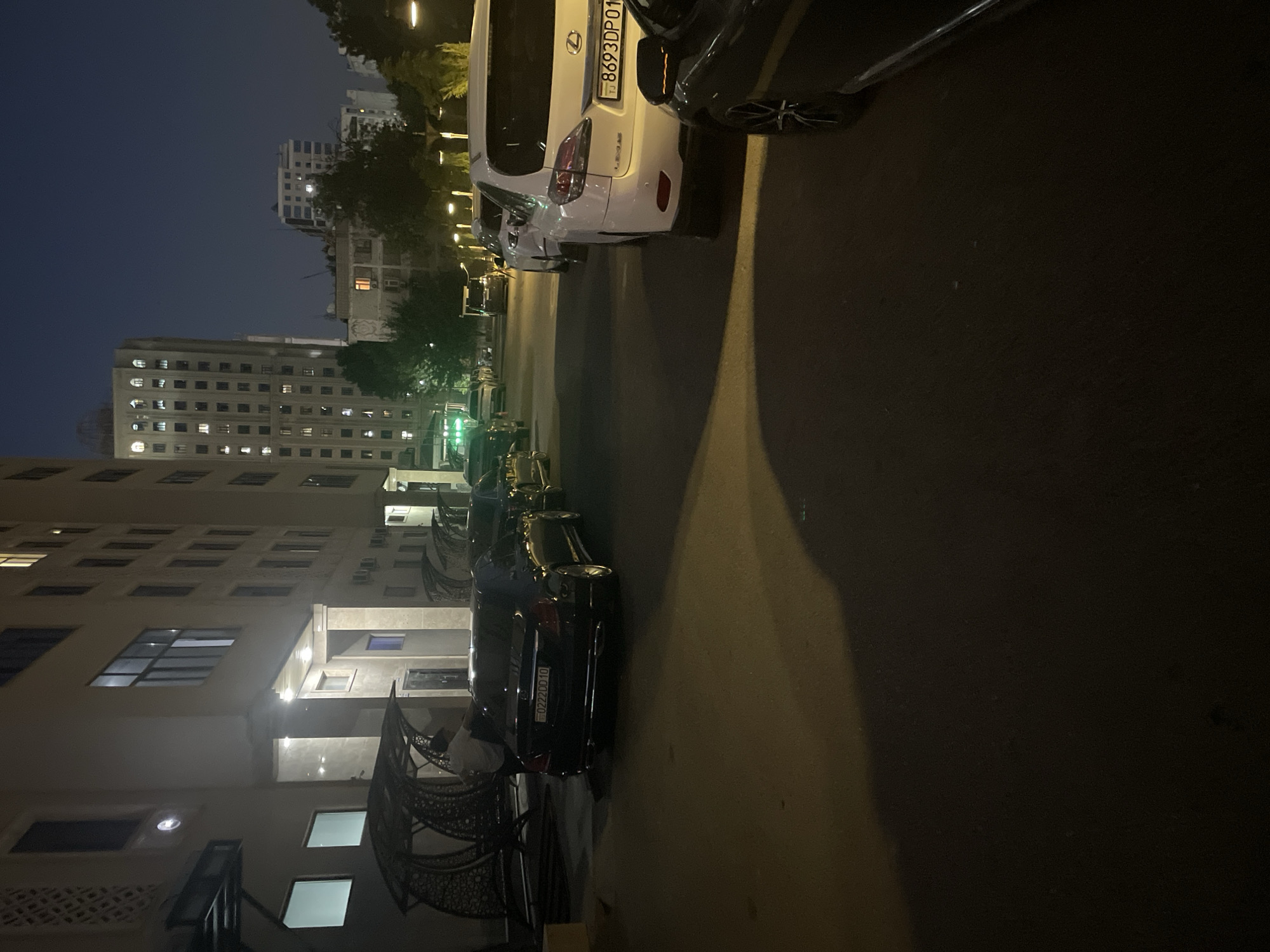 A nighttime exterior shot captures a street scene in Dushanbe, Tajikistan. The foreground features a dark-colored sedan parked on the right, partially obscured by a shadow. To its left, a white Lexus SUV is visible, with its license plate reading "TJ 8693DP01". Further down the street, more cars are parked and lined up. The background reveals the architecture of the city, with illuminated buildings and streetlights contributing to the nocturnal ambiance.