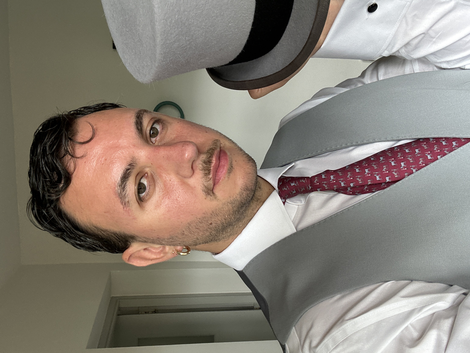An indoor, chest-up portrait captures a male subject in formal wear, situated in what appears to be a residential or private setting in London, United Kingdom. The man, with short, dark, slicked-back hair and a light beard and mustache, looks directly at the viewer. His left earlobe features a gold hoop earring. He is dressed in a white long-sleeved dress shirt, over which a grey waistcoat is worn. A maroon tie with a repeating pattern of small white animal figures, possibly horses, is visible against the shirt. To the right of the primary subject, partially obscured, a hand holds a light grey top hat with a black band. Another section of a white shirt with a visible black cufflink is also present on the right side of the frame. The background consists of plain, light-colored walls, with a partially open doorway visible on the left and a faint circular, teal object on the wall to the right.