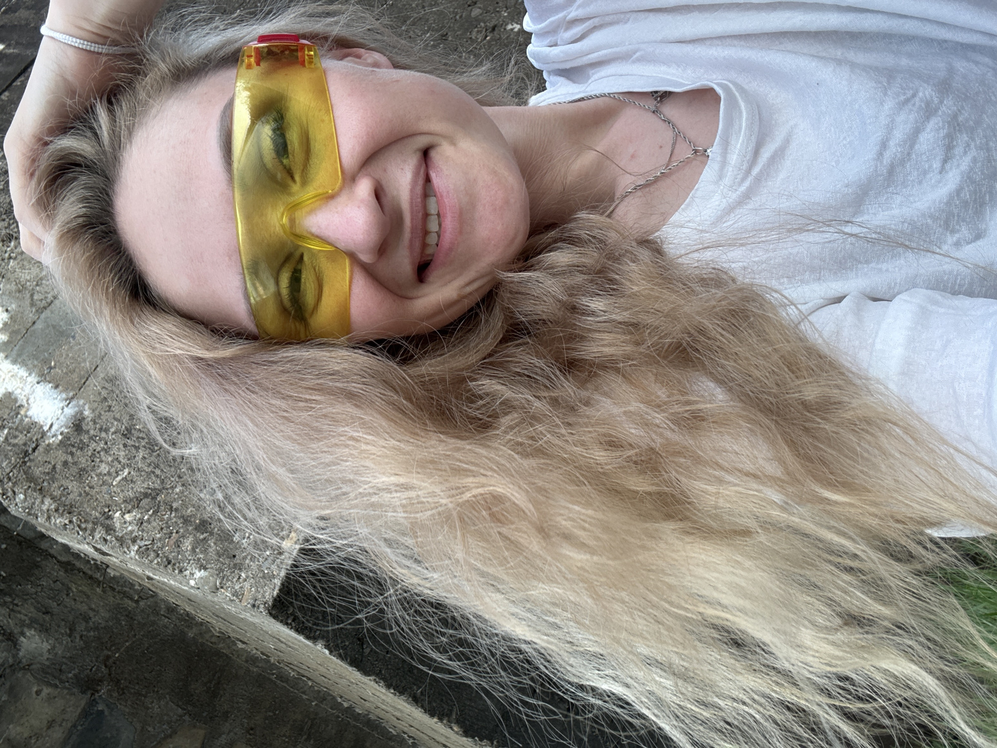 A woman with long blonde hair, wearing a white t-shirt and yellow sunglasses, is lying on her side on what appears to be a concrete surface. She is smiling and holding her head with one hand. A silver necklace is visible around her neck. The location cannot be determined based on the given image.