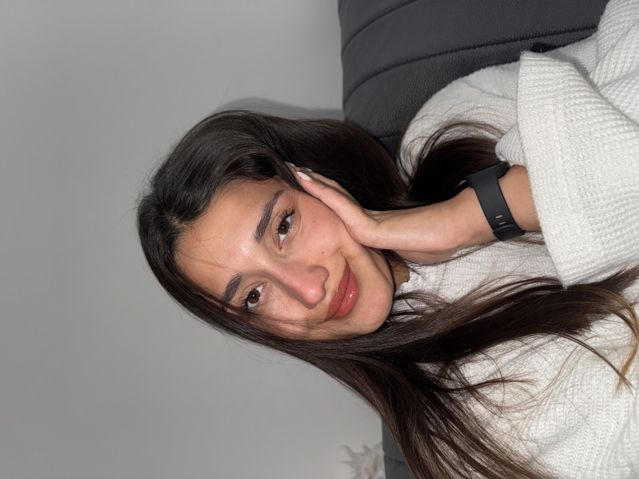 A young adult female is depicted in a close-up, indoor portrait. She possesses long, dark brown hair, brown eyes, and a slight smile while looking directly at the camera. Her left hand is positioned against her left cheek, displaying white nail polish, and a dark grey or black digital smartwatch with a silicone band is present on her left wrist. She is dressed in a white, textured knit sweater. To her right, a segment of dark grey upholstered furniture with vertical stitching is visible. The background consists of a plain light grey wall, with a blurred, light-colored object partially visible behind her left shoulder. The specific location within the city of Budva, Montenegro, is not discernible from the visual information provided in the image.