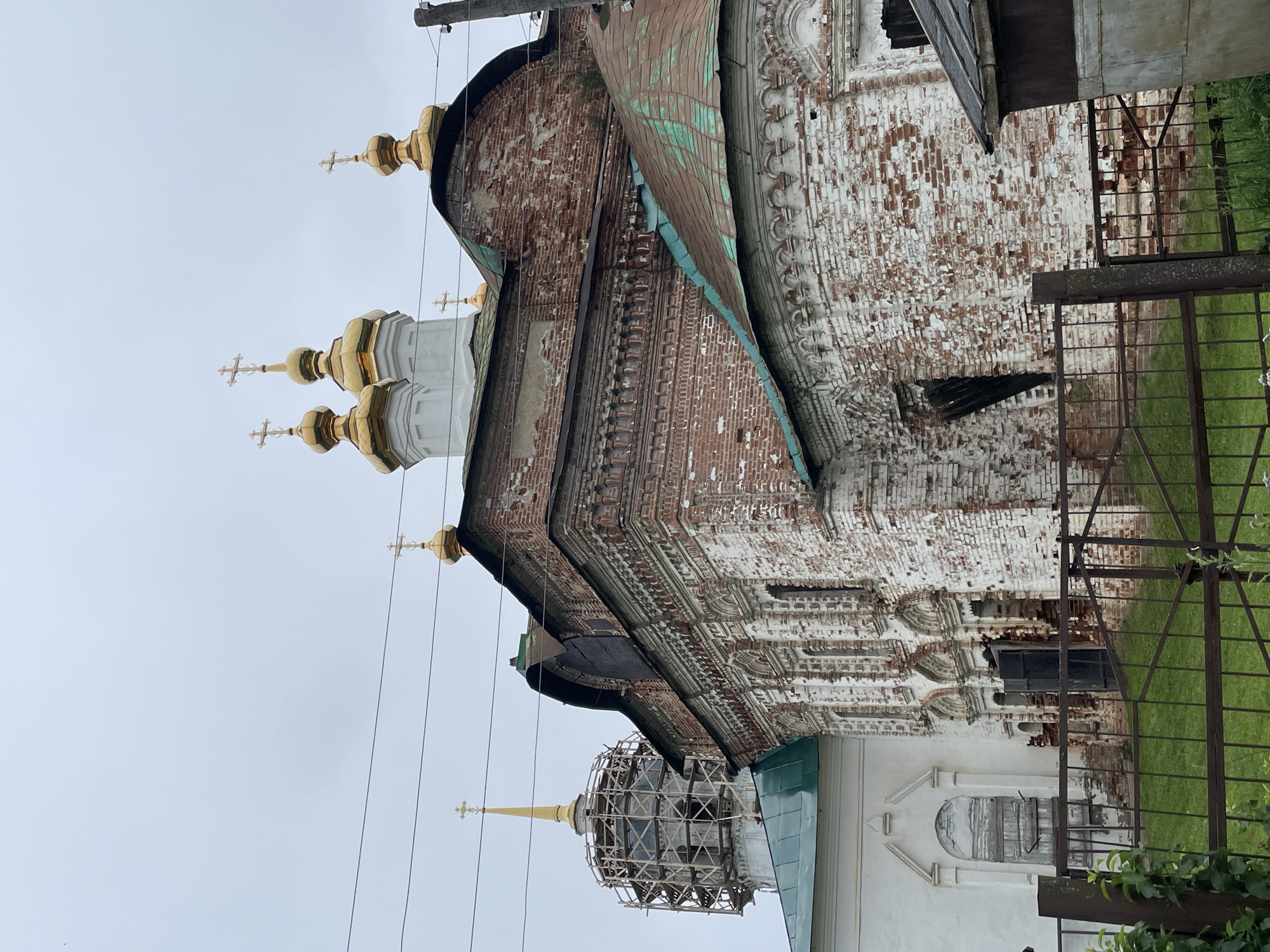 The image shows the exterior of an old, weathered, brick church in Kstinin, Russia. The church features multiple golden domes topped with crosses, a characteristic of Orthodox Christian architecture. The brickwork is a mix of red and white, with significant signs of age and decay, including peeling paint and exposed brick. Some sections of the church appear to be under repair, with scaffolding visible around a tall, slender dome. The sky is overcast, creating a muted lighting condition. In the foreground, a rusty metal fence runs across the scene, with green grass visible behind it. A small, nondescript structure with a slanted roof is partially visible on the right side of the image. The overall impression is one of historical significance and a sense of ongoing preservation efforts for the ancient religious site.