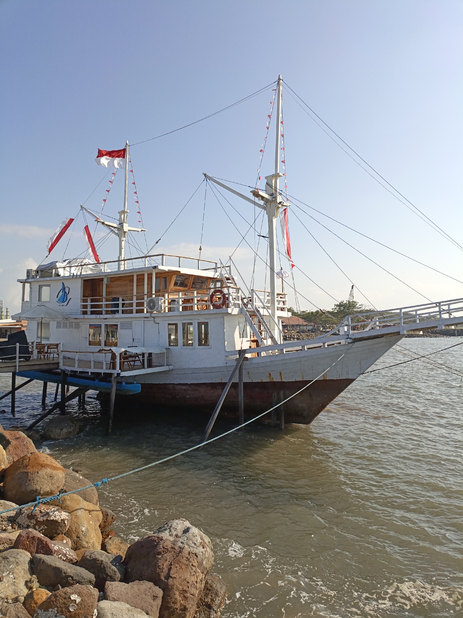 The image shows a white wooden ship docked next to a rocky shoreline under a clear blue sky. The ship, named "PINISI" on its hull, features multiple decks and is adorned with Indonesian flags and small red and white pennant flags, suggesting a celebratory occasion or national holiday. A gangway extends from the shore to the ship, allowing access. The ship is moored with thick ropes to the rocky embankment. The water surrounding the ship appears calm with small ripples. In the background, there are some buildings and trees, hinting at a coastal settlement. The overall scene captures a tranquil maritime setting in Bulukumba, Indonesia, a region renowned for its traditional boat-building heritage.